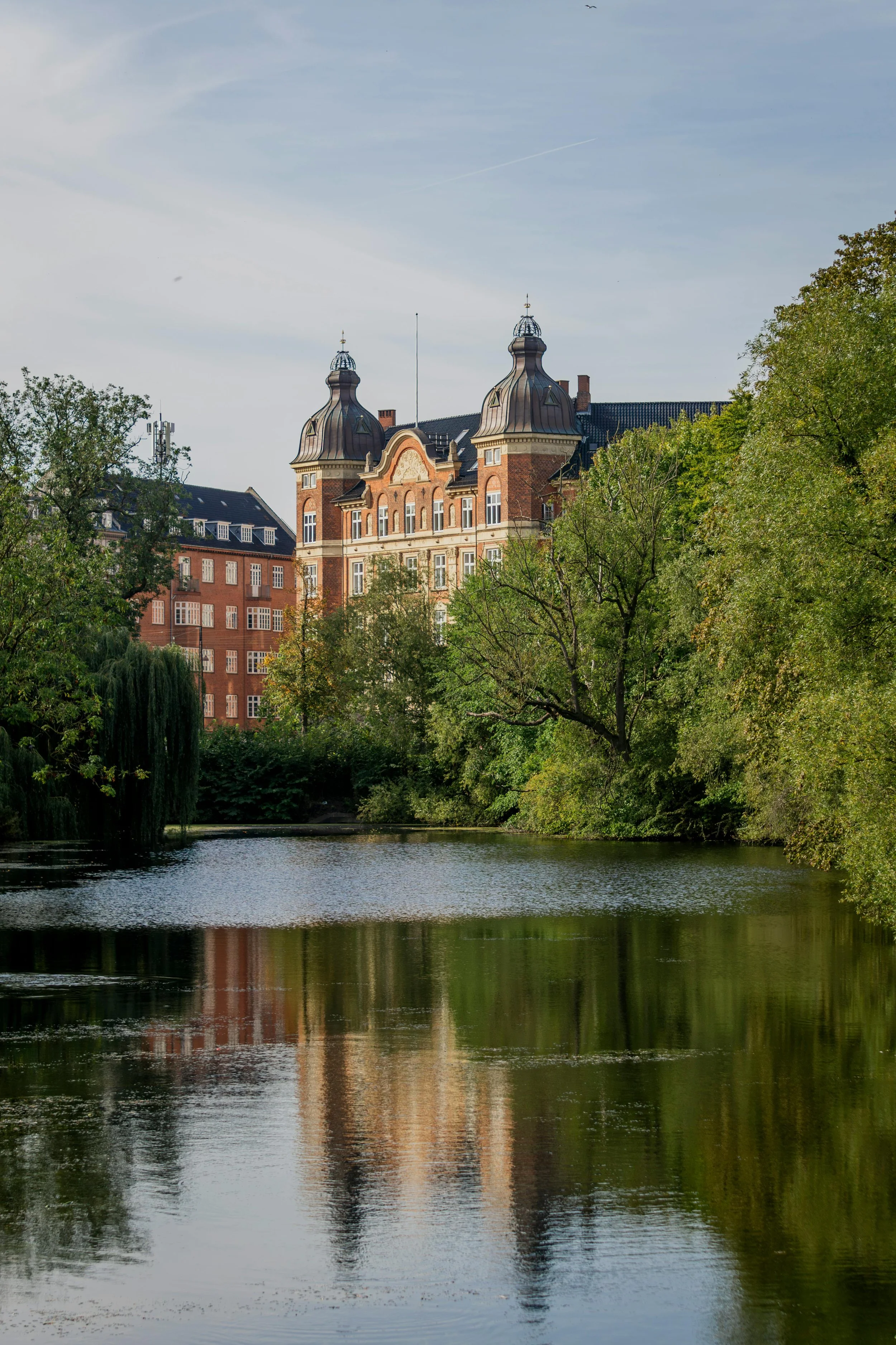 Office in a historic brick building with ornate towers seen above green trees, reflected in a calm river.