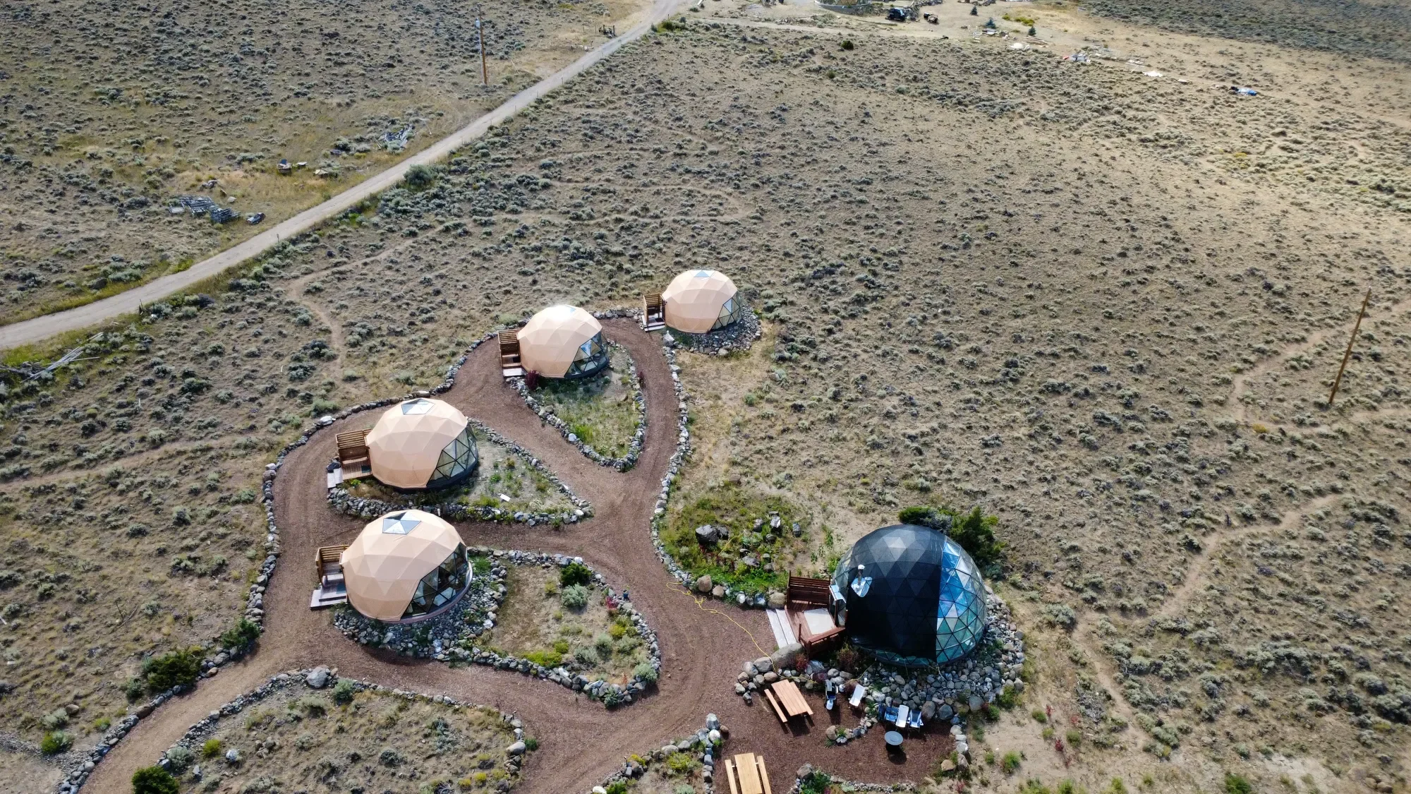 Aerial view of five geodesic dome cabins arranged in a line at Pure Heart Retreat in Wapiti Valley, WY. Each dome is surrounded by gravel and plants, with pathways connecting them. The setting is open with sparse vegetation and dirt roads nearby.