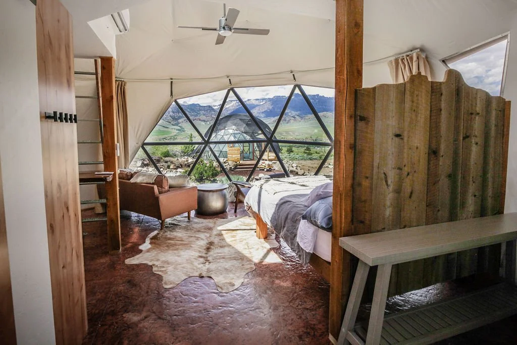 Interior of a rustic glamping tent with a view of mountains and greenery outside. The room has a bed, a cowhide rug, a sitting area with a chair and side table, and a wooden partition.