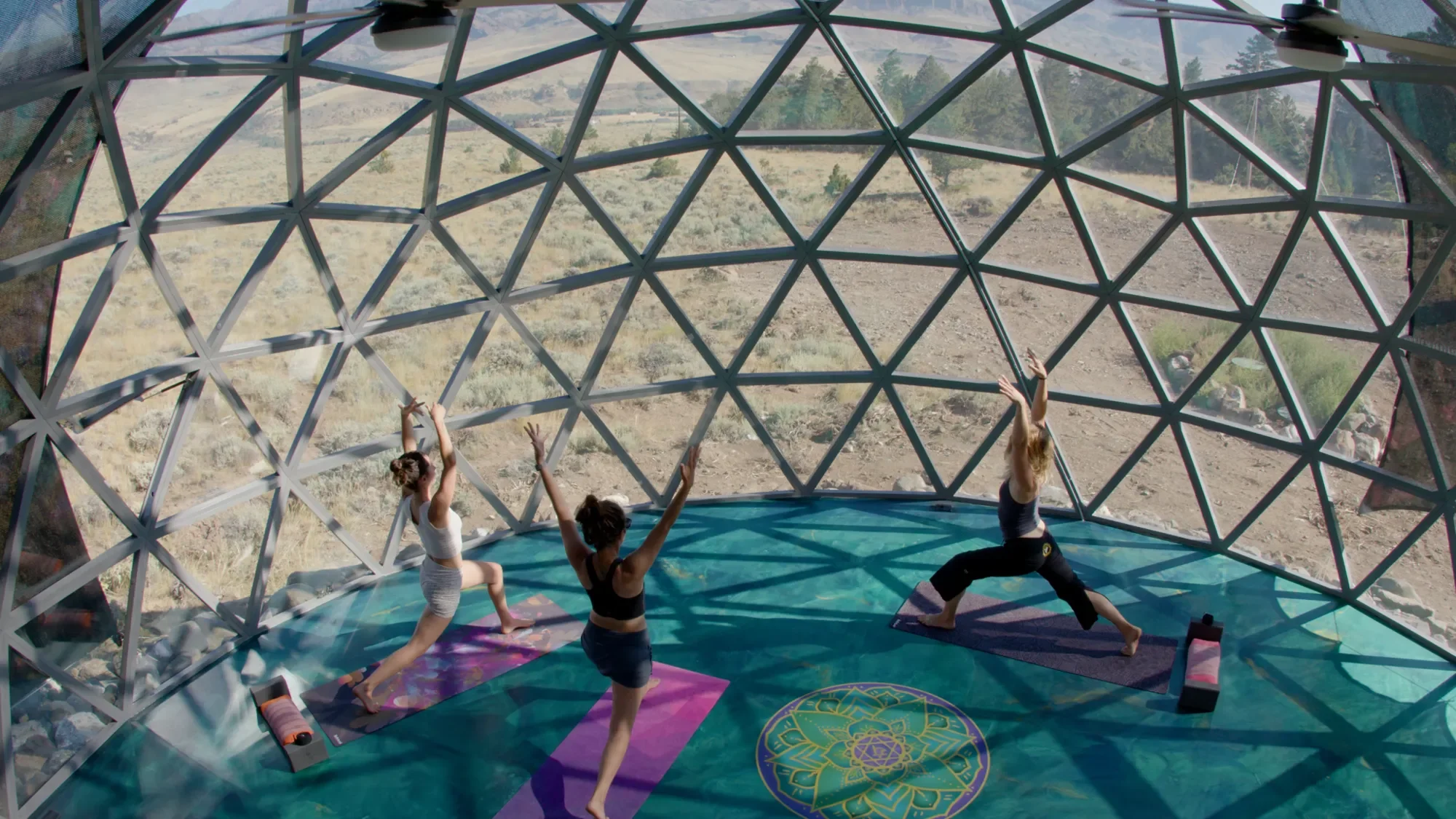 Three women practicing yoga inside a geodesic dome with large windows overlooking a desert landscape.