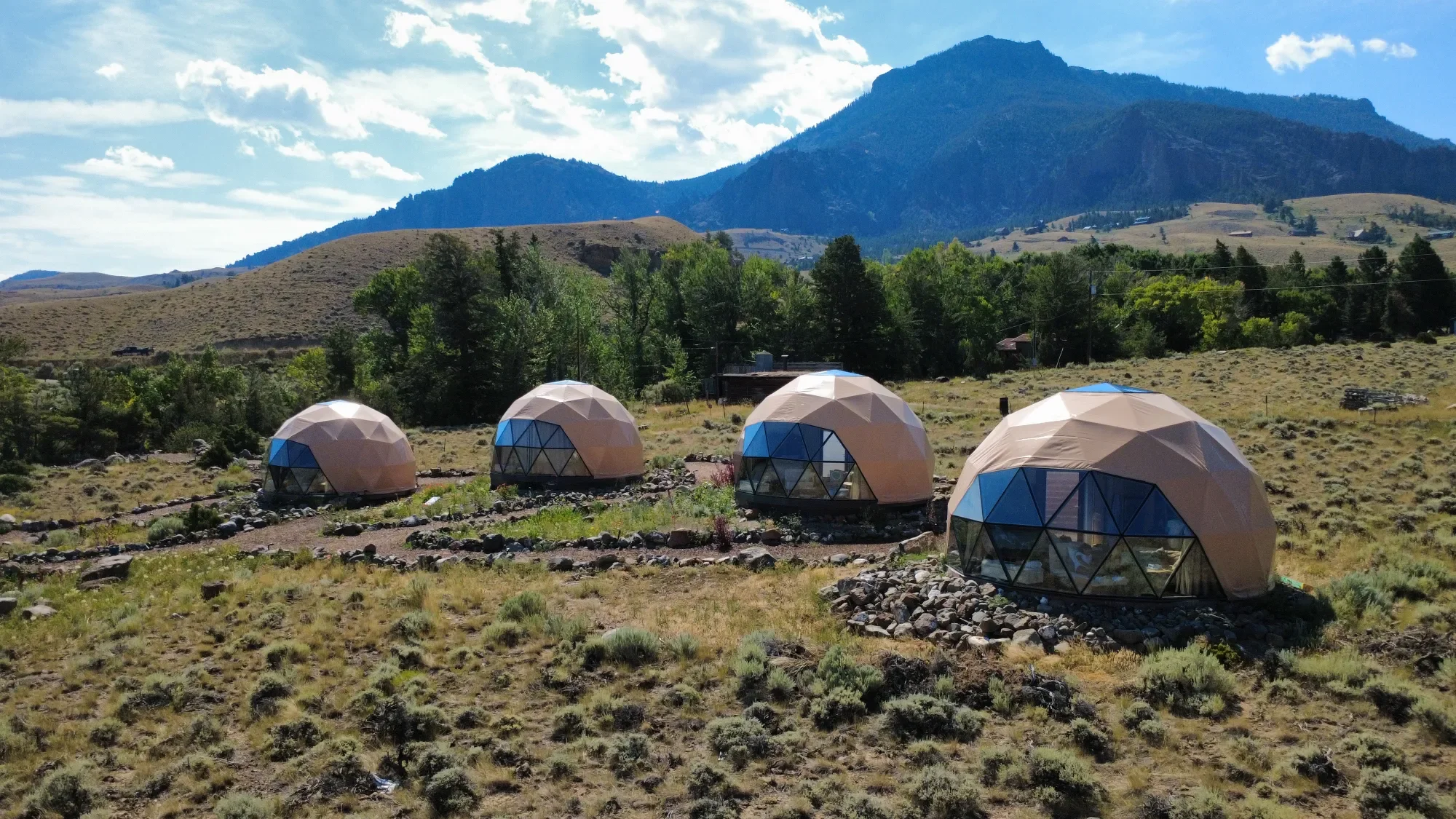 Four geodesic dome tents with large glass windows are set up on a grassy, rocky plain with Wapiti Valley, Wyoming's mountains and trees in the background under a partly cloudy sky.