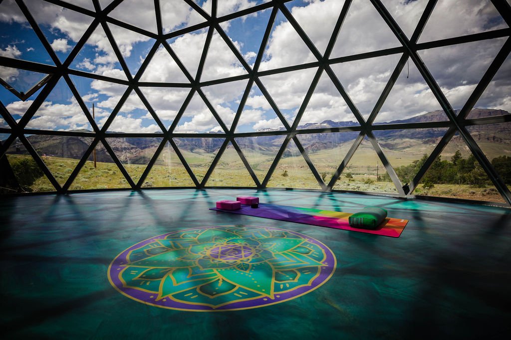 Interior of a geodesic dome with a yoga mat and blocks on the floor, overlooking a mountainous landscape under a partly cloudy sky.