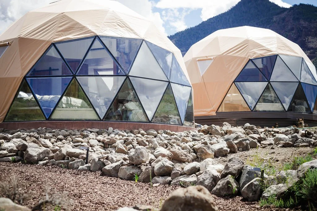 Two geodesic dome structures with glass panels and beige fabric coverings, set on rocky ground in a mountainous area under a partly cloudy sky.