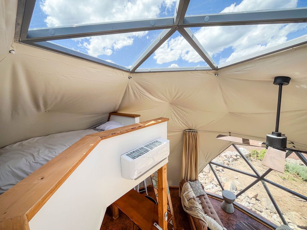 Interior of a geodesic glamping dome with a bed, window, and skylight showing blue sky with clouds. Located in Wapiti Valley, Wyoming at Pure Heart Retreat.