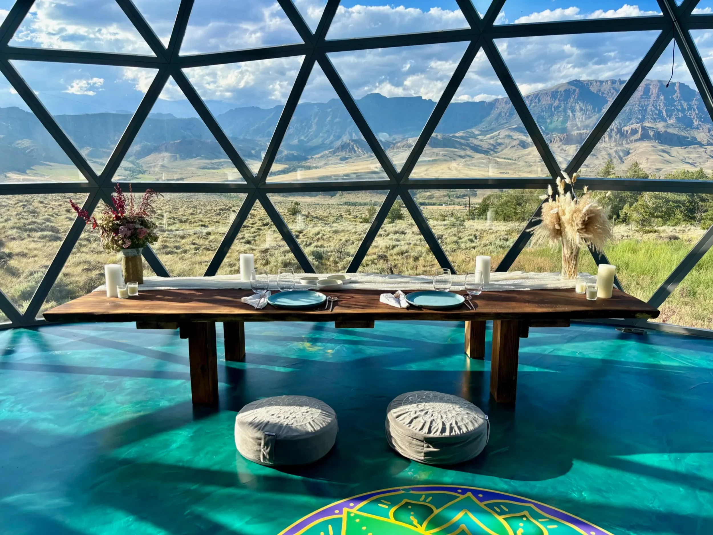 Indoor dining table with two cushions, decorated with flowers, candles, and tableware, overlooking a mountainous landscape through geometric window panels at Pure Heart Retreat in Wapiti, WY. Private retreats and dining services are available.