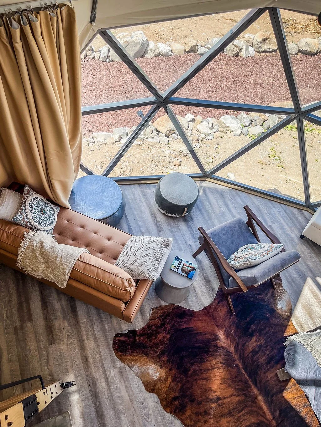 Interior of a geodesic dome room with a tan leather sofa, gray armchair, small side table, and natural fiber pillows. There are two round ottomans near a large geometric window showing a rocky outdoor landscape. A cowhide rug covers part of the wooden floor.