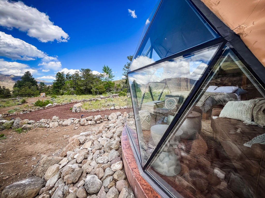 Interior of a modern dome rental with large glass windows reflecting the outside scenery of a rural landscape, with a bed and seating area visible through the glass. Located in Wapiti Valley, Wyoming at Pure Heart Retreat.