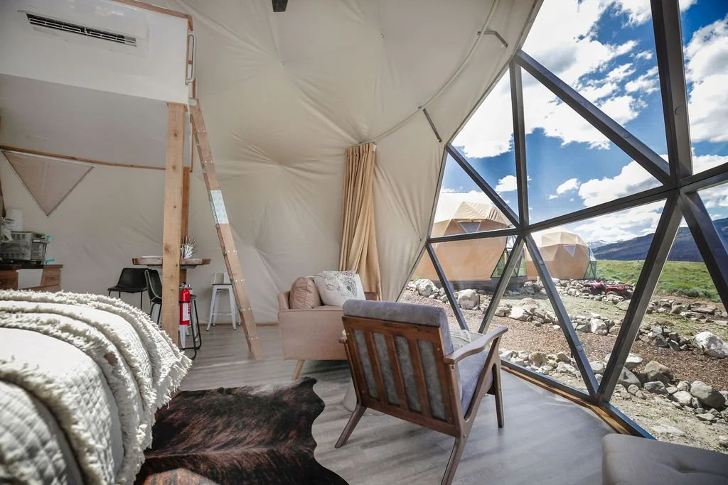 Inside a modern yurt with large geometric window panels, view of mountains and sky, cozy furniture including a chair, sofa, and bed, with a ladder and small kitchen area.