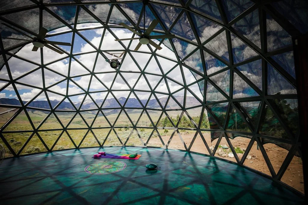 Interior view of a geodesic dome with large glass panels, ceiling fans, and a view of a scenic desert landscape with mountains in the distance.