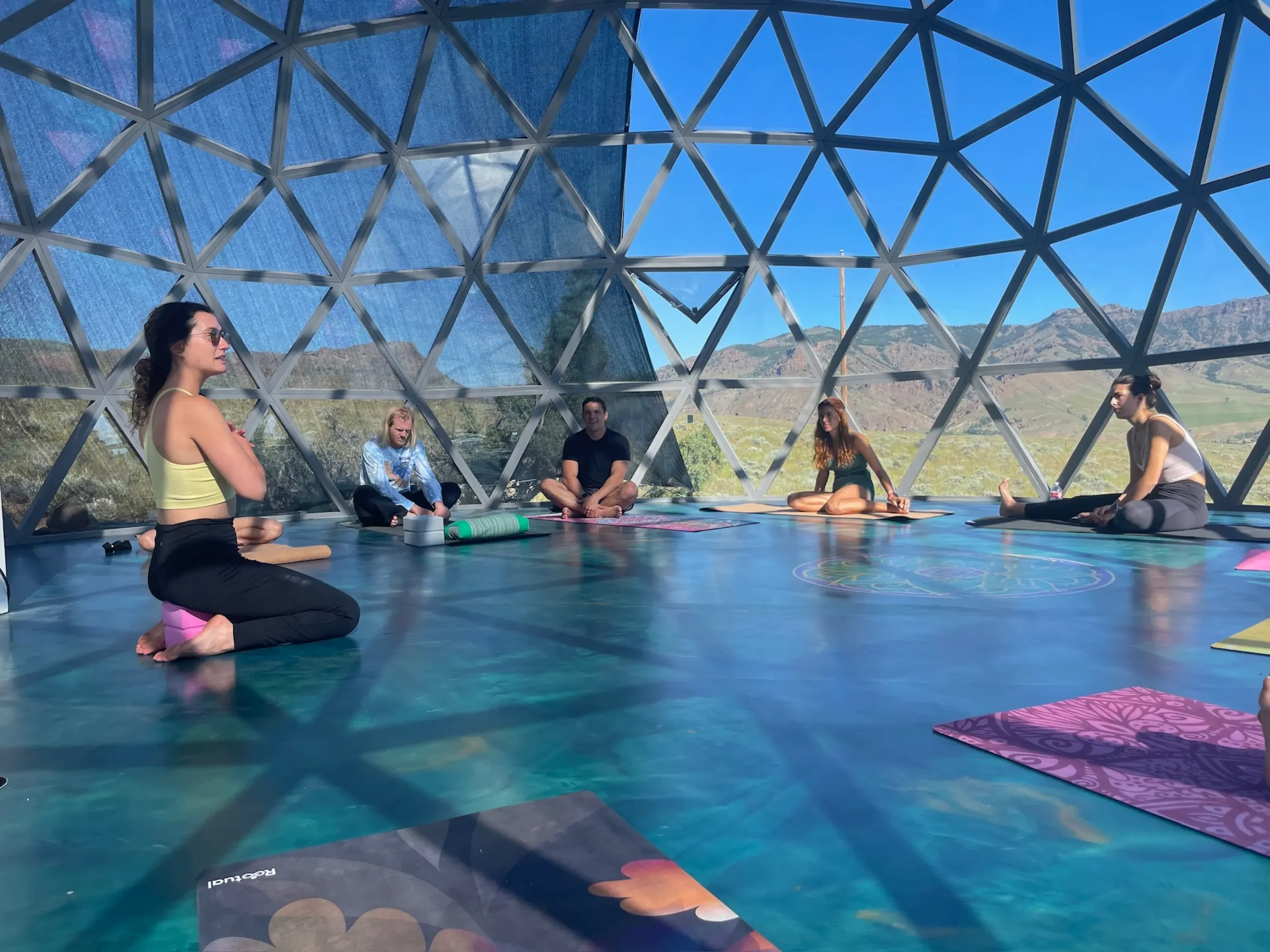 Inside a geodesic dome with a view of mountains and a partly cloudy sky. The floor has a colorful mandala design, and there is yoga equipment including a purple mat with pink blocks and a green bolster.