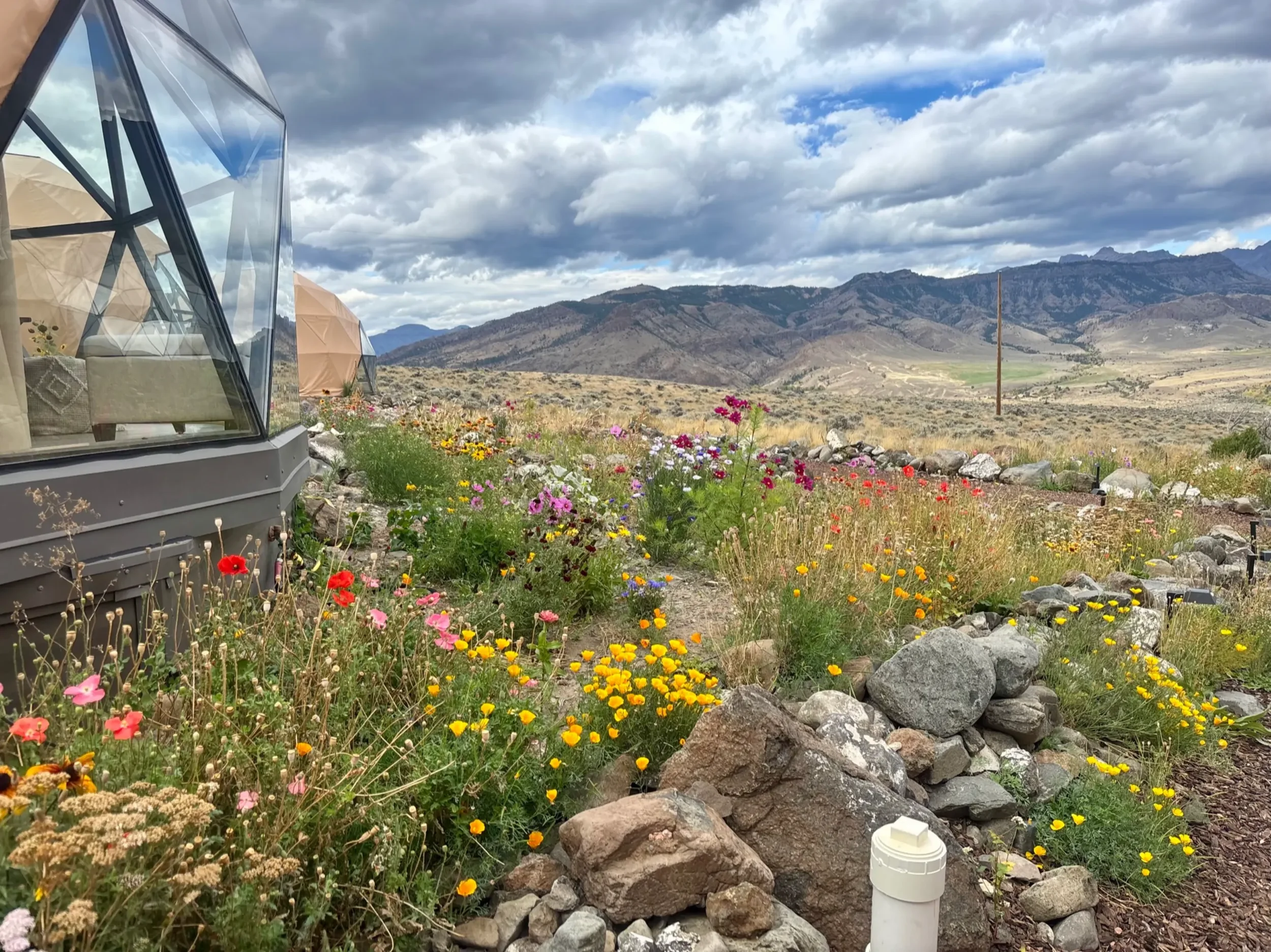 Three geodesic dome structures in a mountainous Wapiti Valley, Wyoming setting with a dirt pathway leading to them, surrounded by rocks and greenery under a partly cloudy sky.