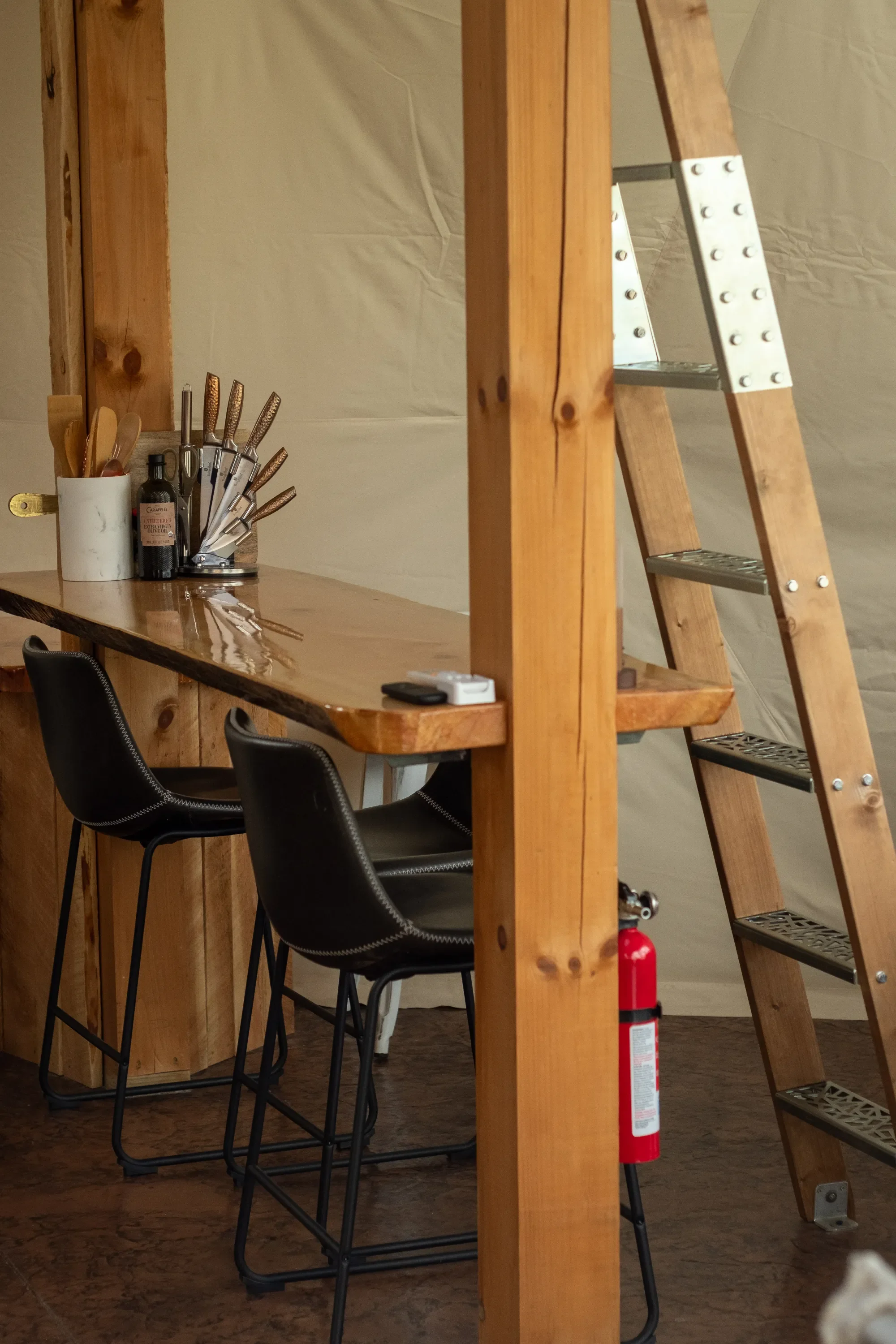 Interior of a wooden bar and kitchen area with a high counter, two black bar stools, a ladder, and various utensils and bottles on the counter. Located in Wapiti Valley, Wyoming at Pure Heart Retreat.