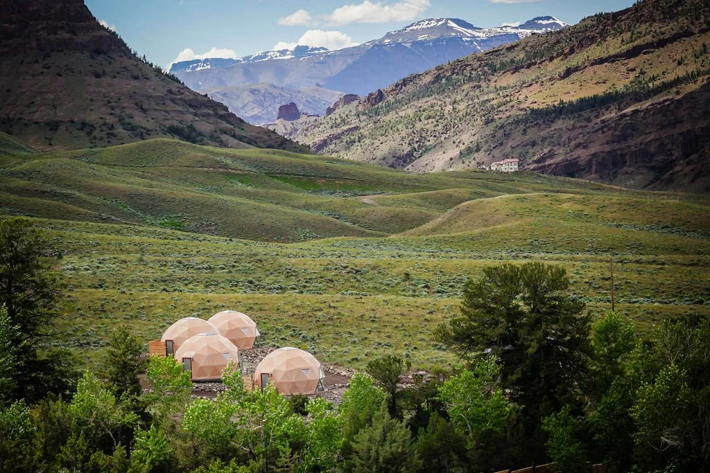 Three geodesic dome structures surrounded by green trees in a mountainous landscape with rolling hills and snow-capped mountains in the distance.