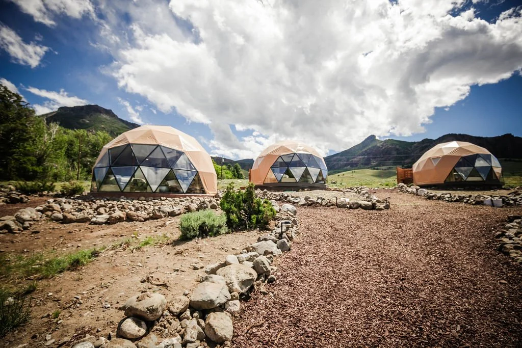 Three geodesic dome structures on rocky terrain with mountains and cloudy sky in the background. Located in Wapiti Valley, Wyoming at Pure Heart Retreat.