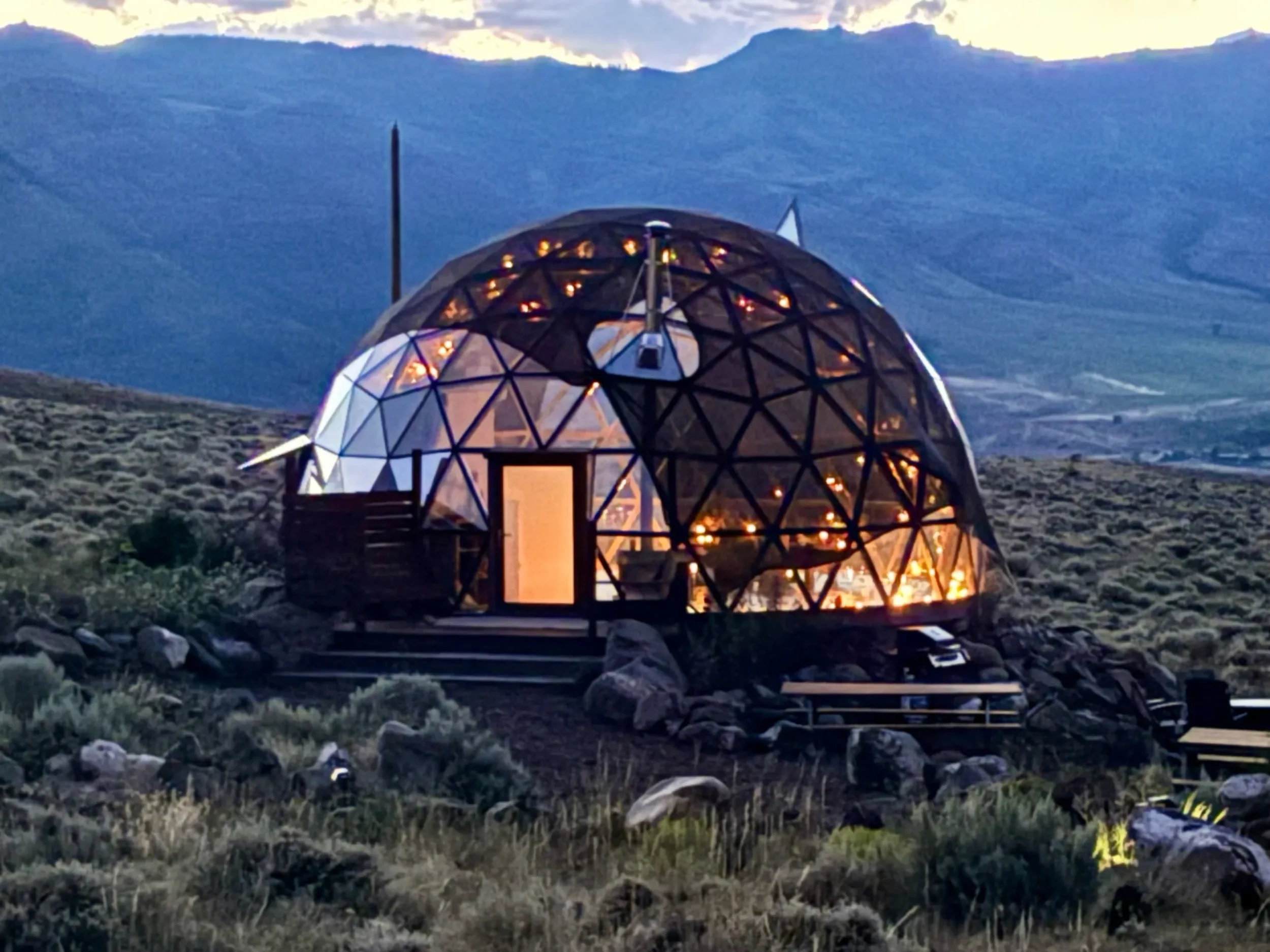 A transparent geodesic dome cabin illuminated with warm lights, set on a rocky, grassy landscape with mountains in the background during twilight at Pure Heart Retreat in Wapiti, WY..