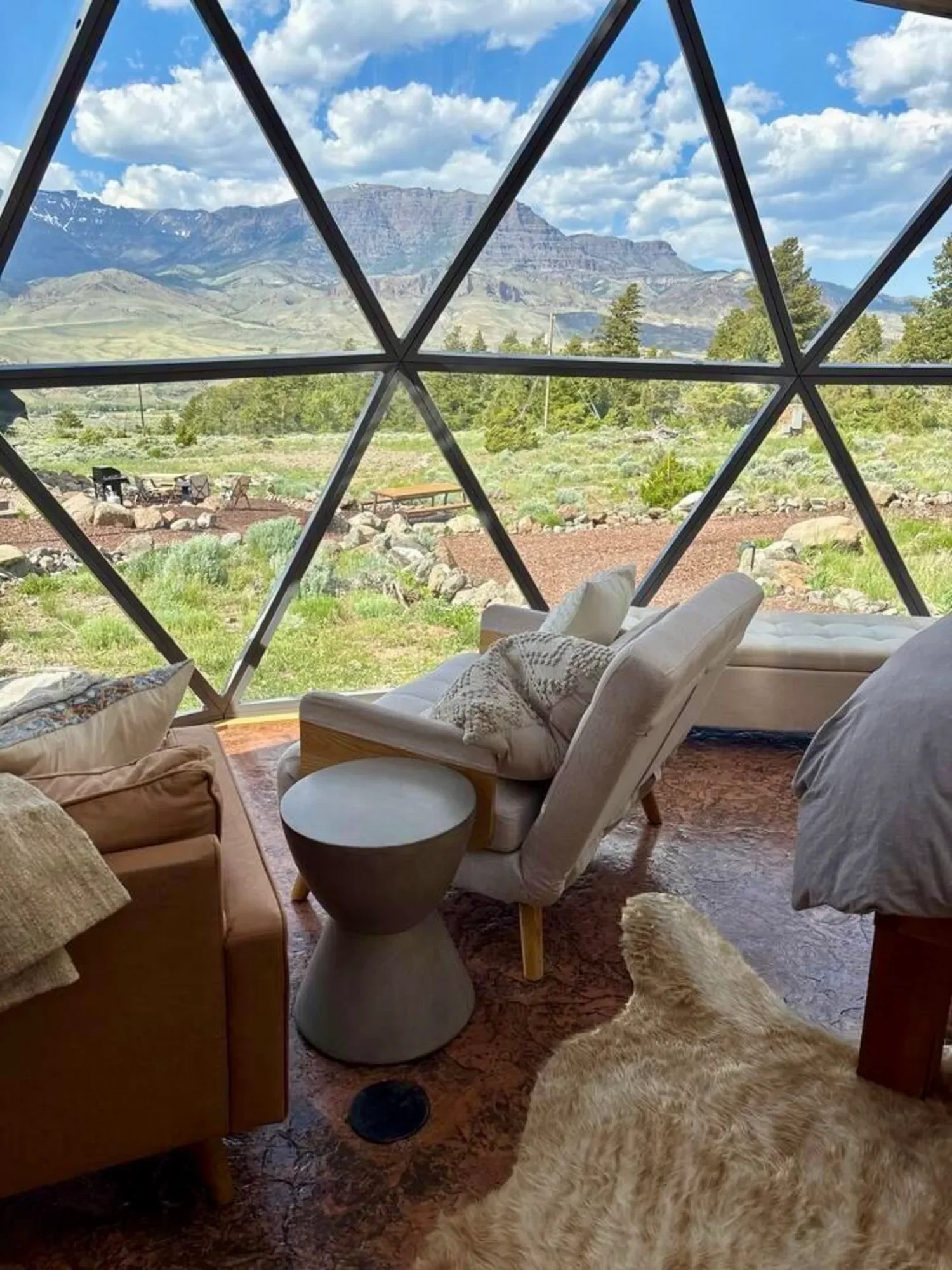 Two beige geodesic dome structures in a natural Wapiti Valley, Wyoming landscape with rocks, greenery, and mountains in the background.