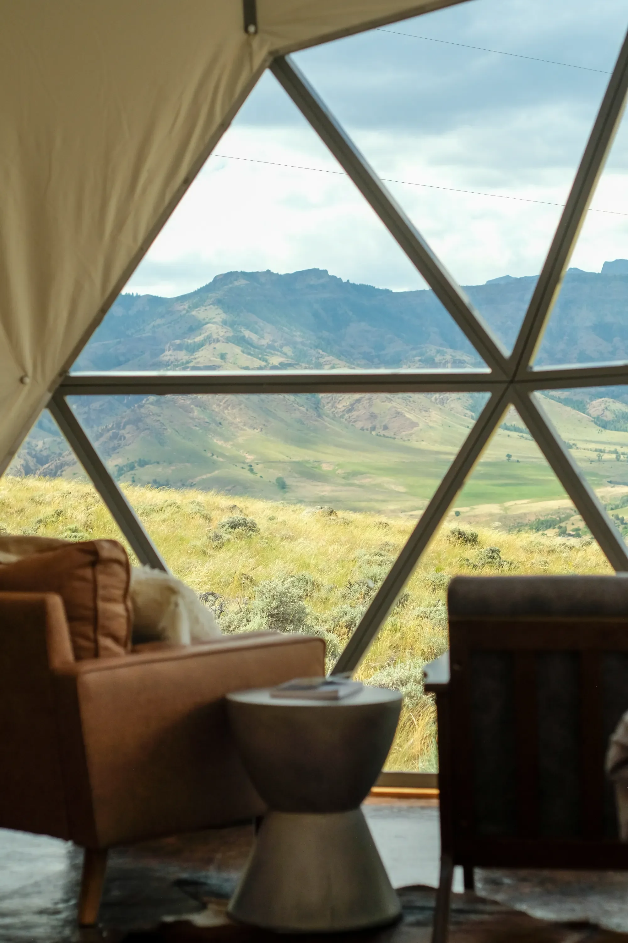 Interior of a yurt with large window showcasing a mountain landscape with grassy hills and mountains under a cloudy sky.