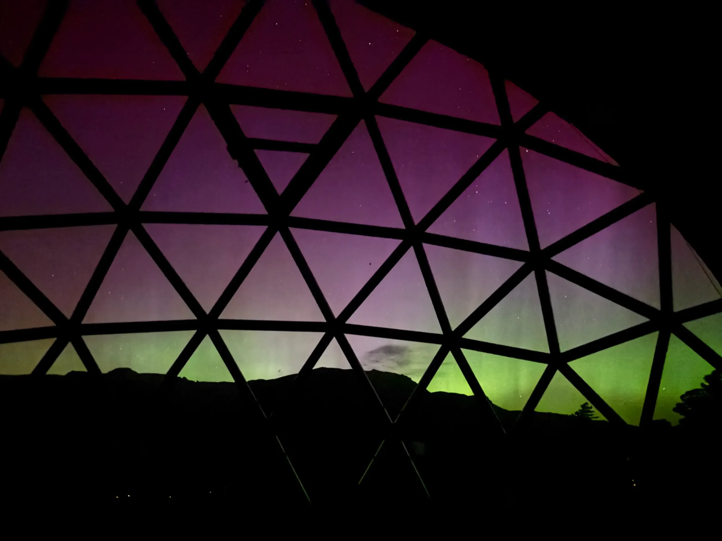 Night sky with purple and green Northern Lights seen through a geometric yoga dome structure at Pure Heart Retreat in Wapiti, WY.
