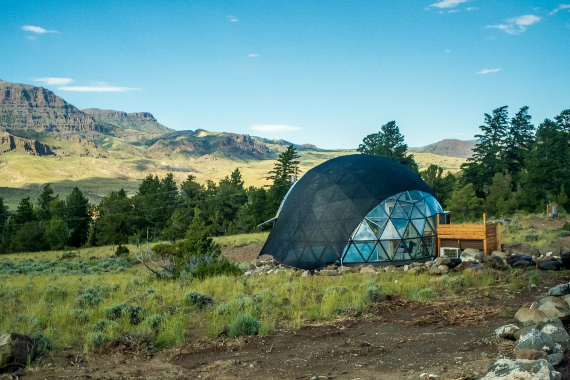 A geodesic dome studio with glass panels and wooden deck outside, situated in a green landscape with mountains and trees in the background. Located in Wapiti Valley, Wyoming at Pure Heart Retreat.