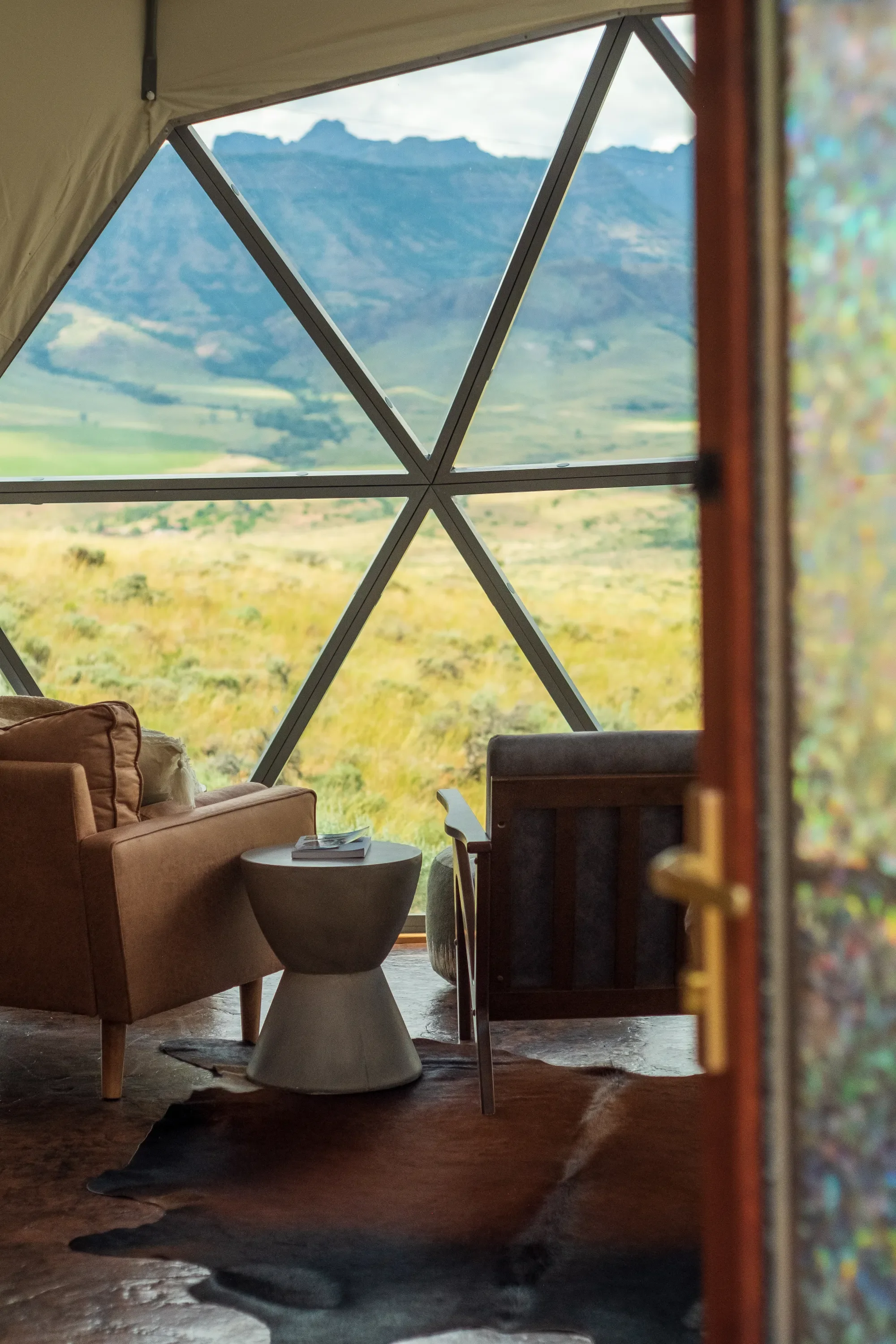 Interior of a cozy living space with a large triangular window showcasing scenic mountains and landscape outside, a brown armchair, a gray armchair, a small side table with magazines, and a cowhide rug on the wooden floor.