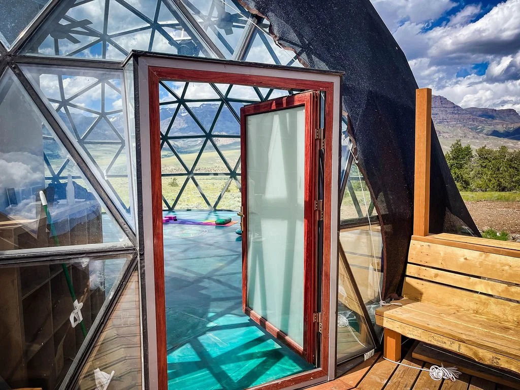 Open wooden and glass door leading into a geodesic yoga dome with a view of mountains and a cloudy sky outside at Pure Heart Retreat in Wapiti, WY.