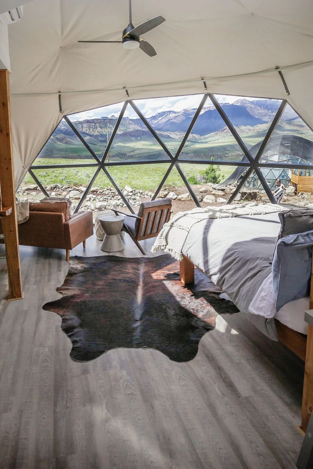 Interior of a yurt with a large geodesic window showing mountains and a landscape outside, furniture including a bed, chairs, a cowhide rug, and a ceiling fan.