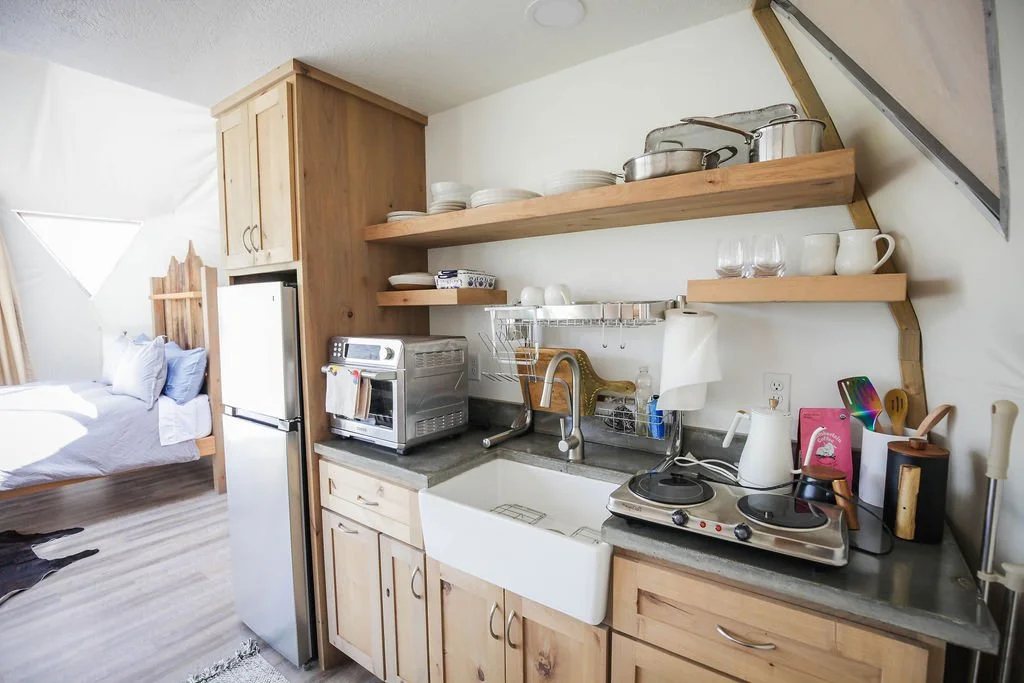 Kitchen with open shelves holding dishes, pots, and glasses, a small sink, a countertop with a toaster oven, electric cooktop, kettle, and utensils, with a bedroom visible in the background.