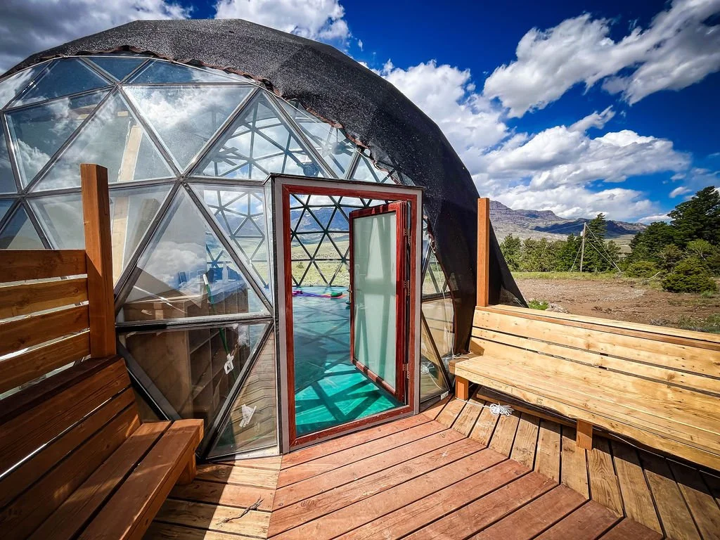 A geodesic dome yoga studio with glass panels and a wooden deck, set in a scenic outdoor area with mountains and trees, under a partly cloudy sky. Located in Wapiti Valley, Wyoming at Pure Heart Retreat.