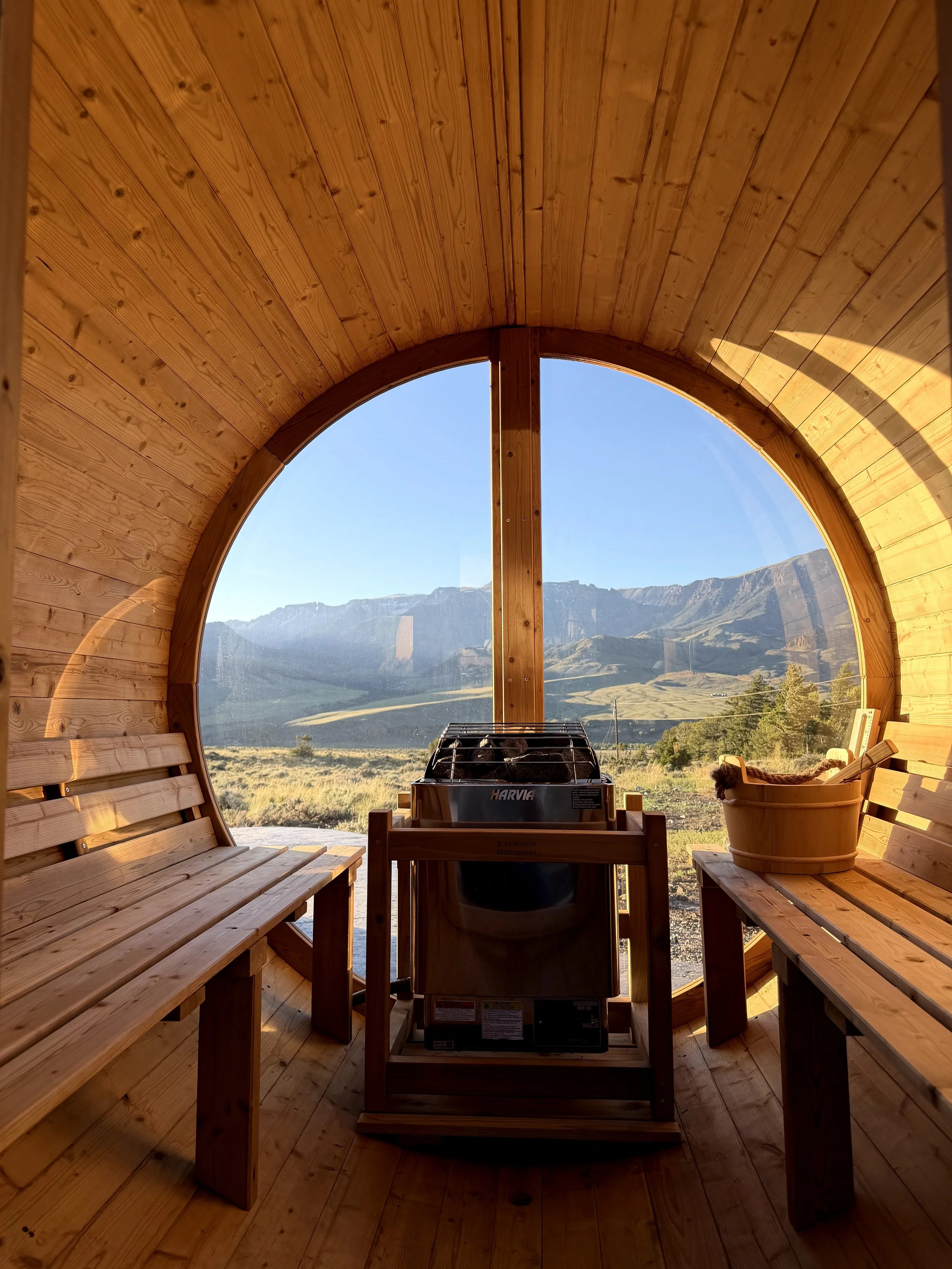 Wooden sauna with open door, situated inside a glass yoga dome with a view of Wapiti Valley, Wyoming outside.