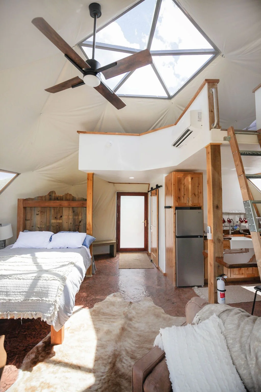 Interior of a cozy, rustic-style glamping dome loft with a bed on the left, a cowhide rug on the floor, a ceiling fan, and a skylight showing blue sky and clouds. Located in Wapiti Valley, Wyoming at Pure Heart Retreat.