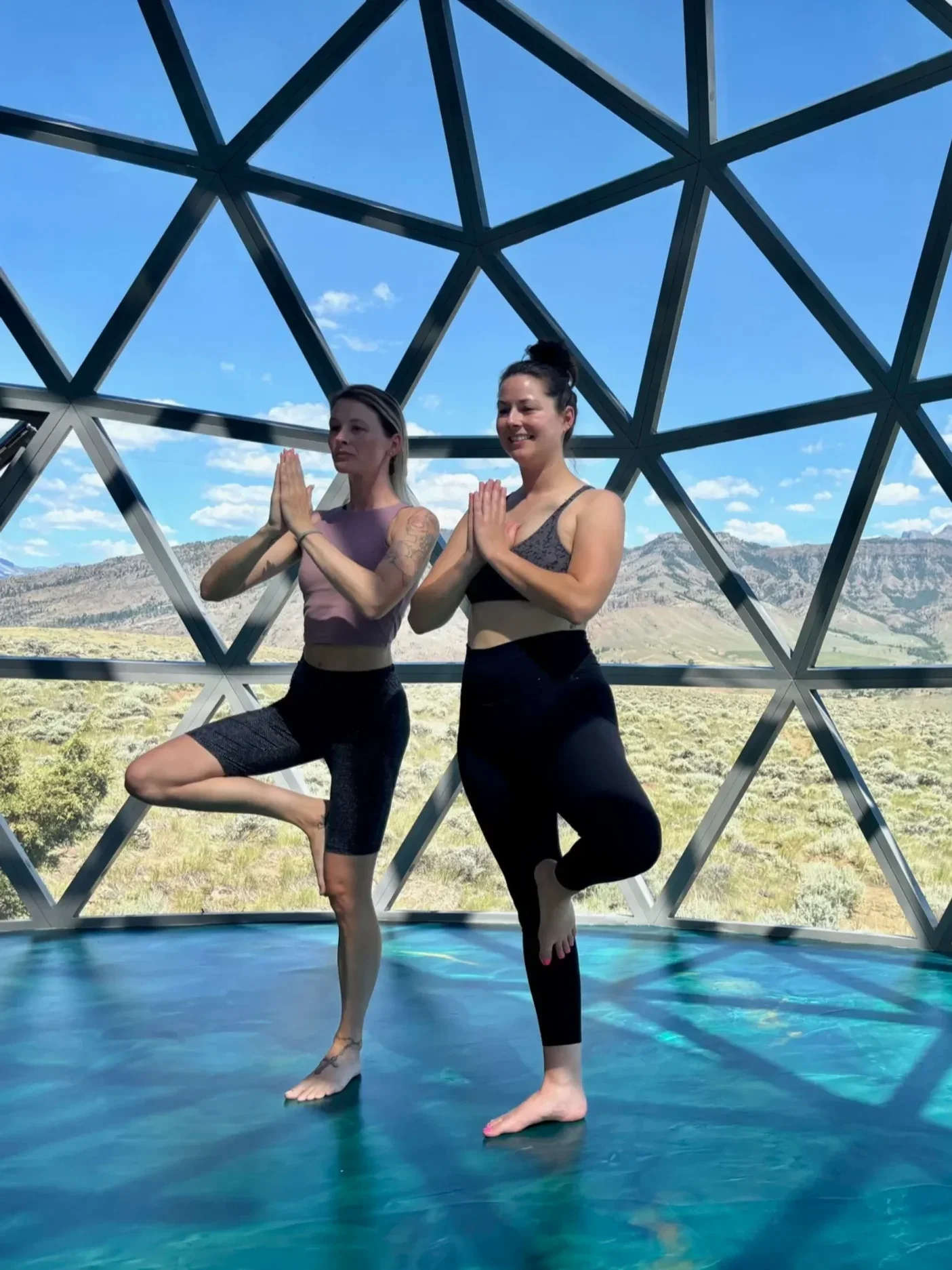 Two women practicing yoga on mats in a geodesic dome at Pure Heart Retreat with snowy mountains in the background.