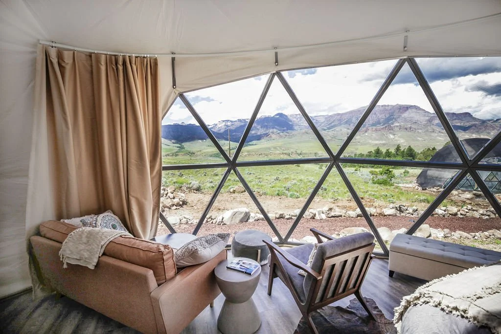 A geodesic dome house with glass walls set in Wapiti Valley, Wyoming's Pure Heart Retreat center with trees and mountains under a partly cloudy sky.