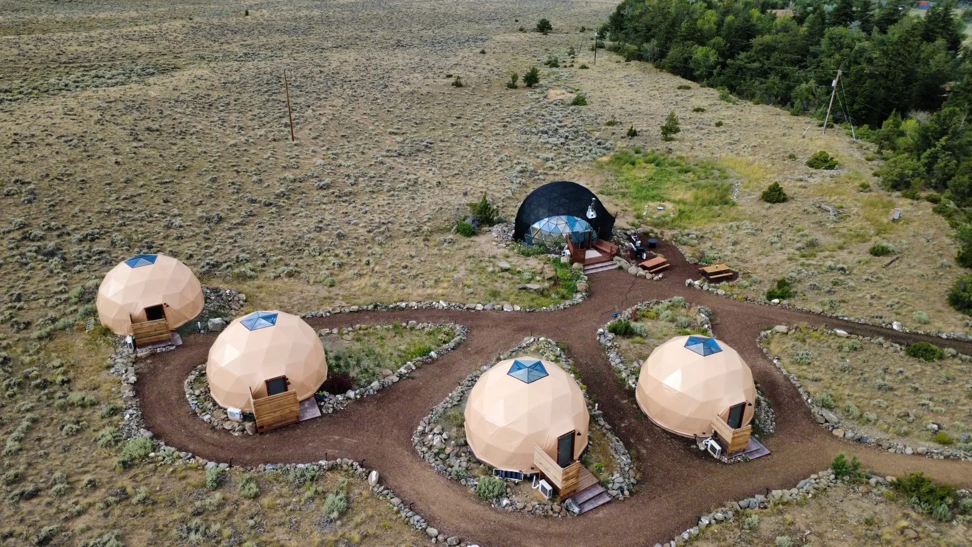 An aerial view of four geodesic dome cabins arranged in a circular pattern on a dirt pathway in a rural, semi-arid landscape with sparse vegetation and a forested area in the background.