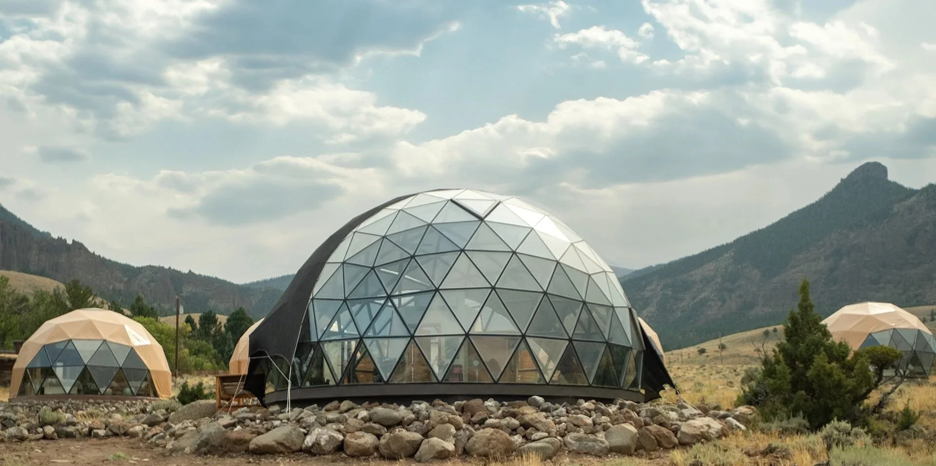 Glass geodesic dome vacation rental situated on the rocky terrain of Wapiti, Valley, WY with mountains and cloudy sky in the background. Located at Pure Heart Retreat.