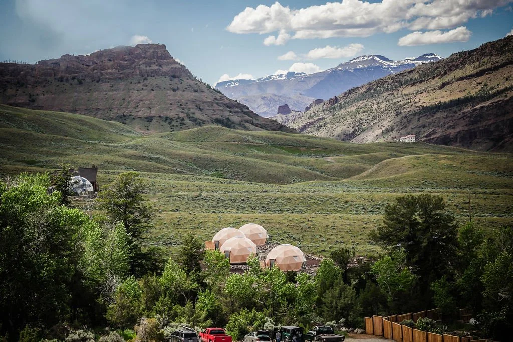 A landscape of rolling green hills and mountains with snow-capped peaks in the distance, some buildings, and several pink geodesic dome structures among trees in the foreground.