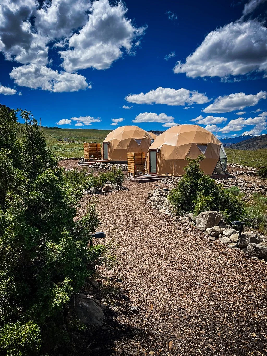 Two geodesic dome cabins on a gravel path in a mountainous landscape under a partly cloudy sky.