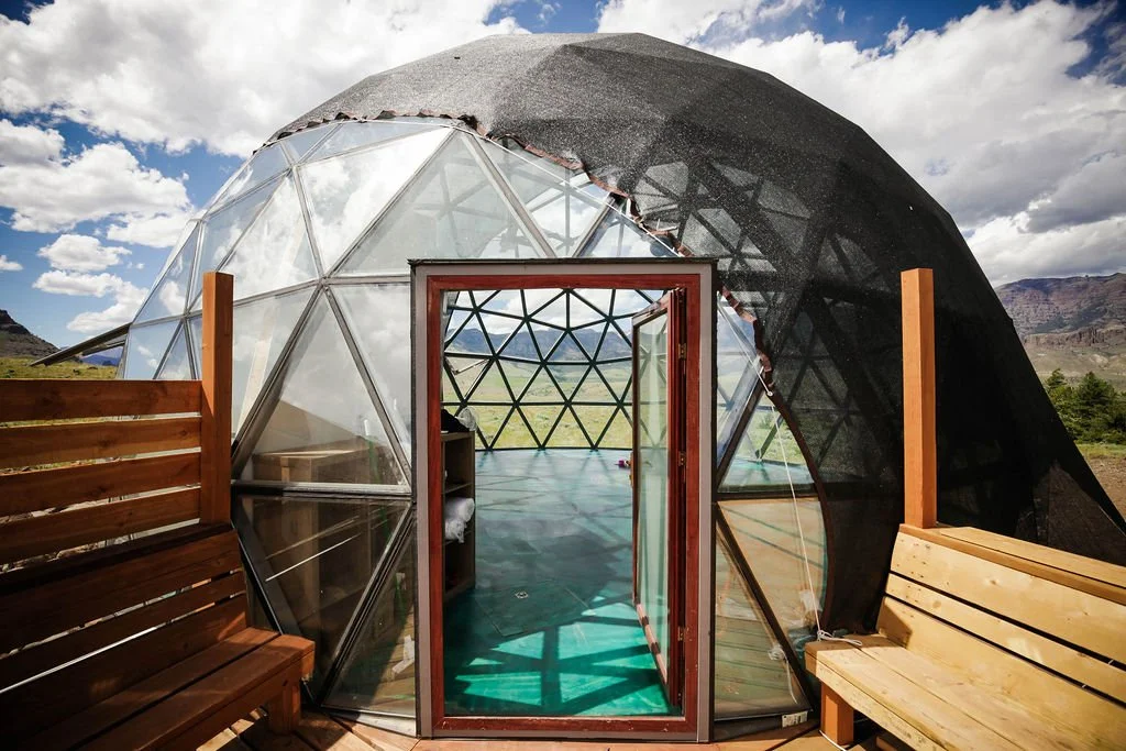 A geodesic dome yoga studio with a glass and wood door, surrounded by wooden benches, located in a rural landscape with mountains in the background.