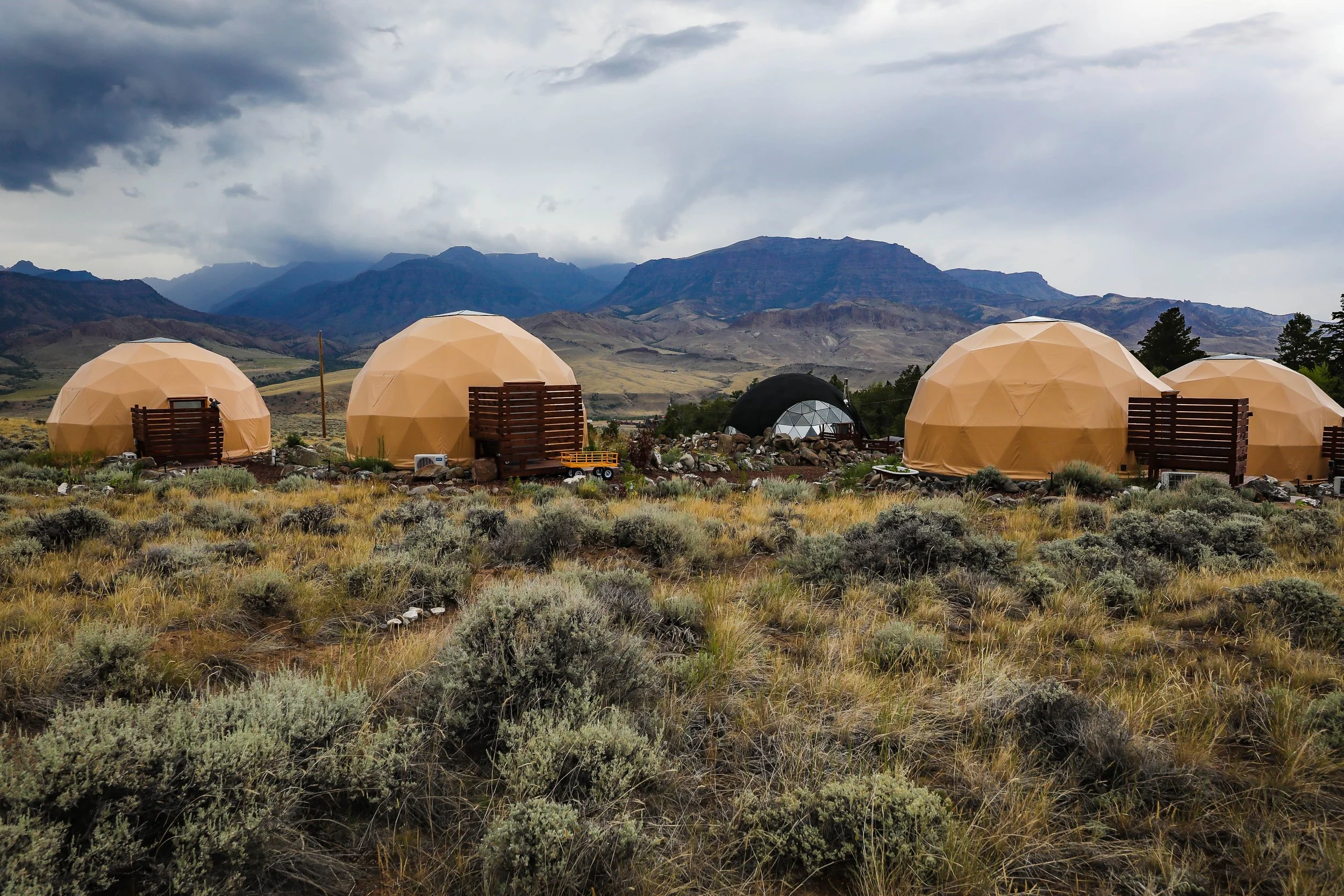 Three geodesic dome structures on rocky terrain with mountains and cloudy sky in the background. Located in Wapiti Valley, Wyoming at Pure Heart Retreat.