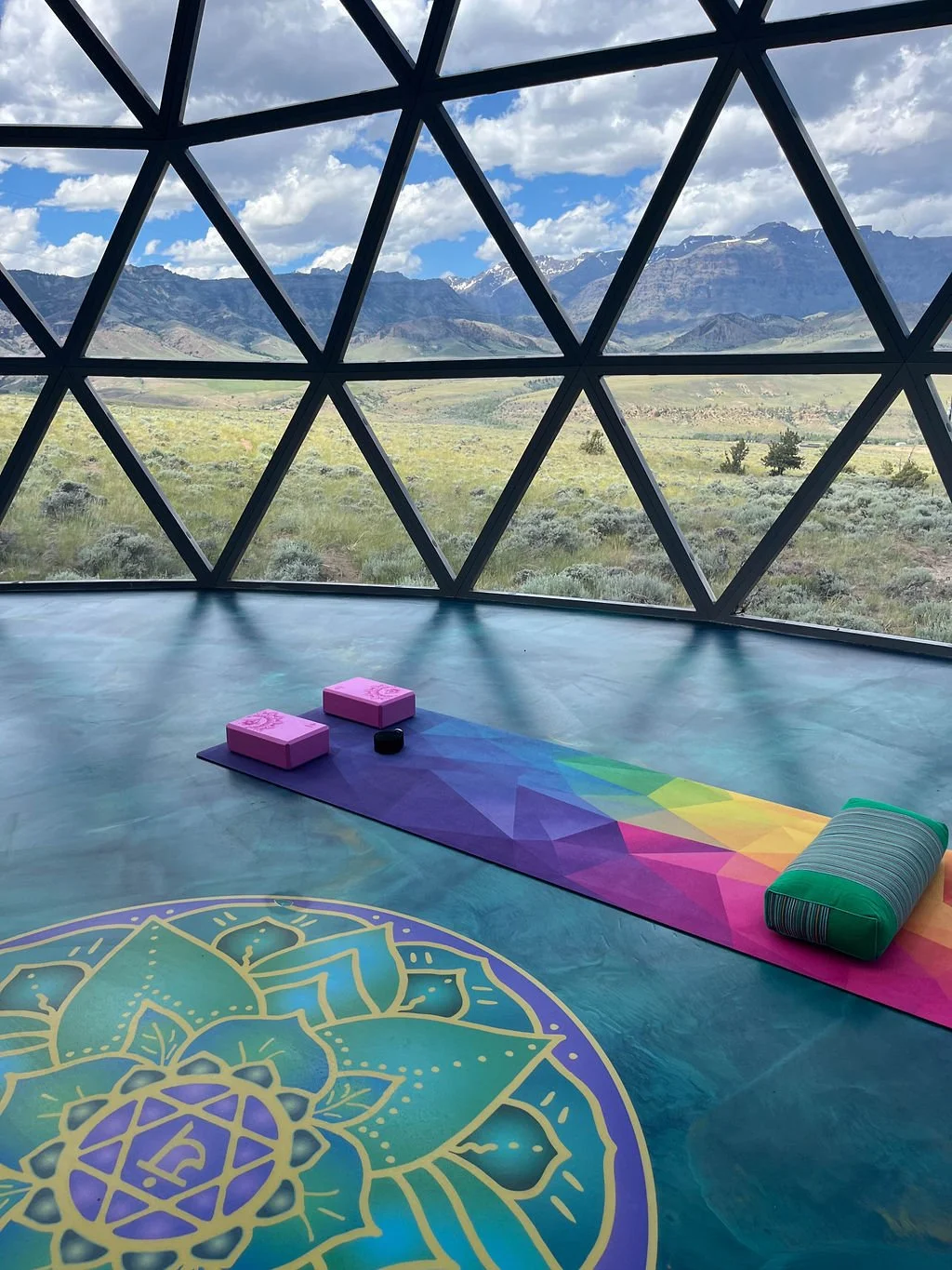 Inside a geodesic dome, a yoga mat with blocks and a bolster on it, with a mountain landscape and blue sky with clouds outside the window.