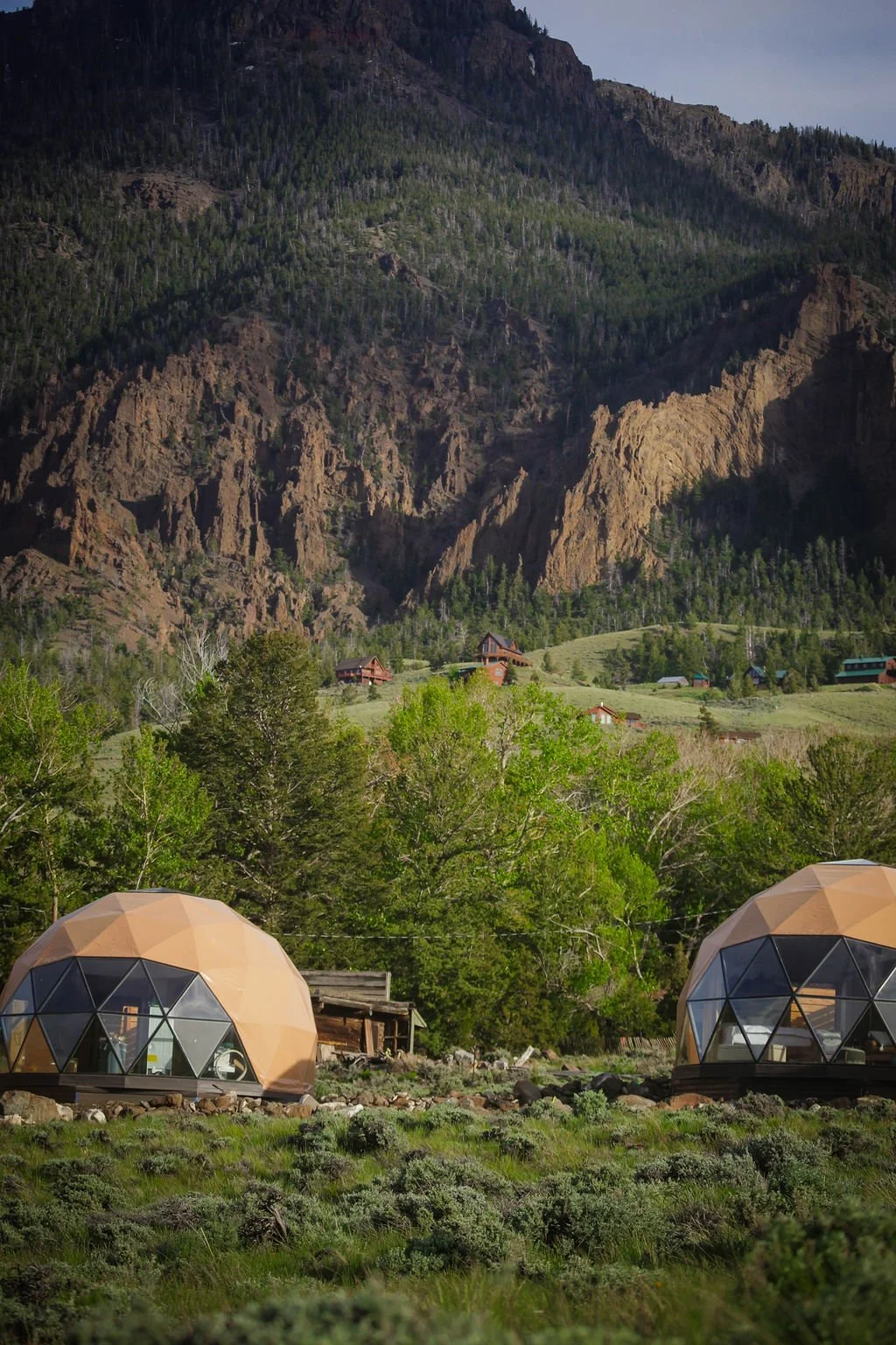 Two geodesic dome structures in a lush green landscape with trees, set against a mountainous backdrop and scattered houses.