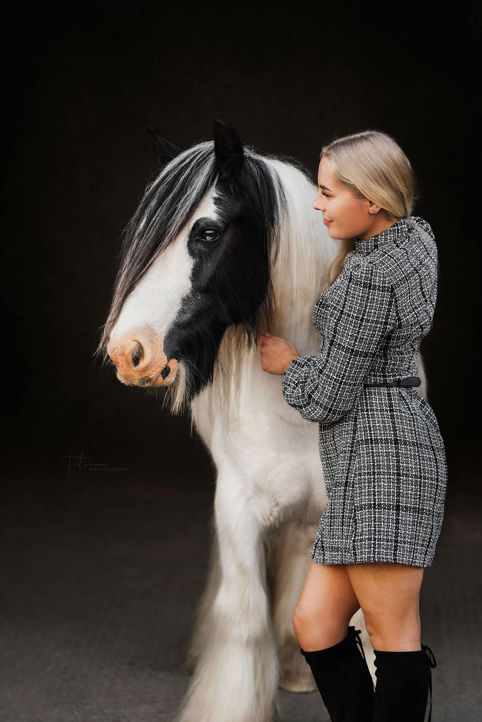 A woman with blonde hair wearing a grey checkered dress and black knee-high boots gently touching a black and white horse against a dark background.