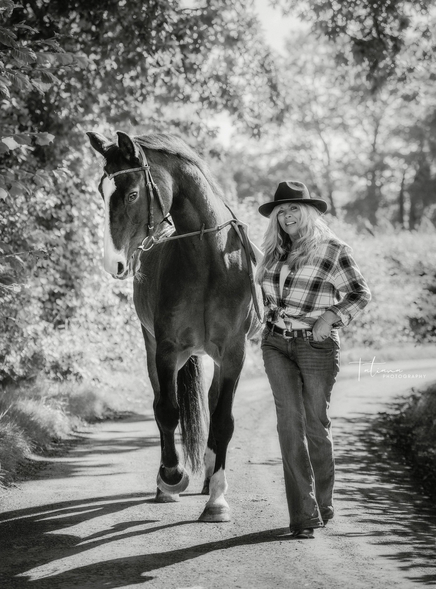 A woman with long hair, wearing a plaid shirt, jeans, and a wide-brimmed hat, standing next to a large horse on a tree-lined dirt path.