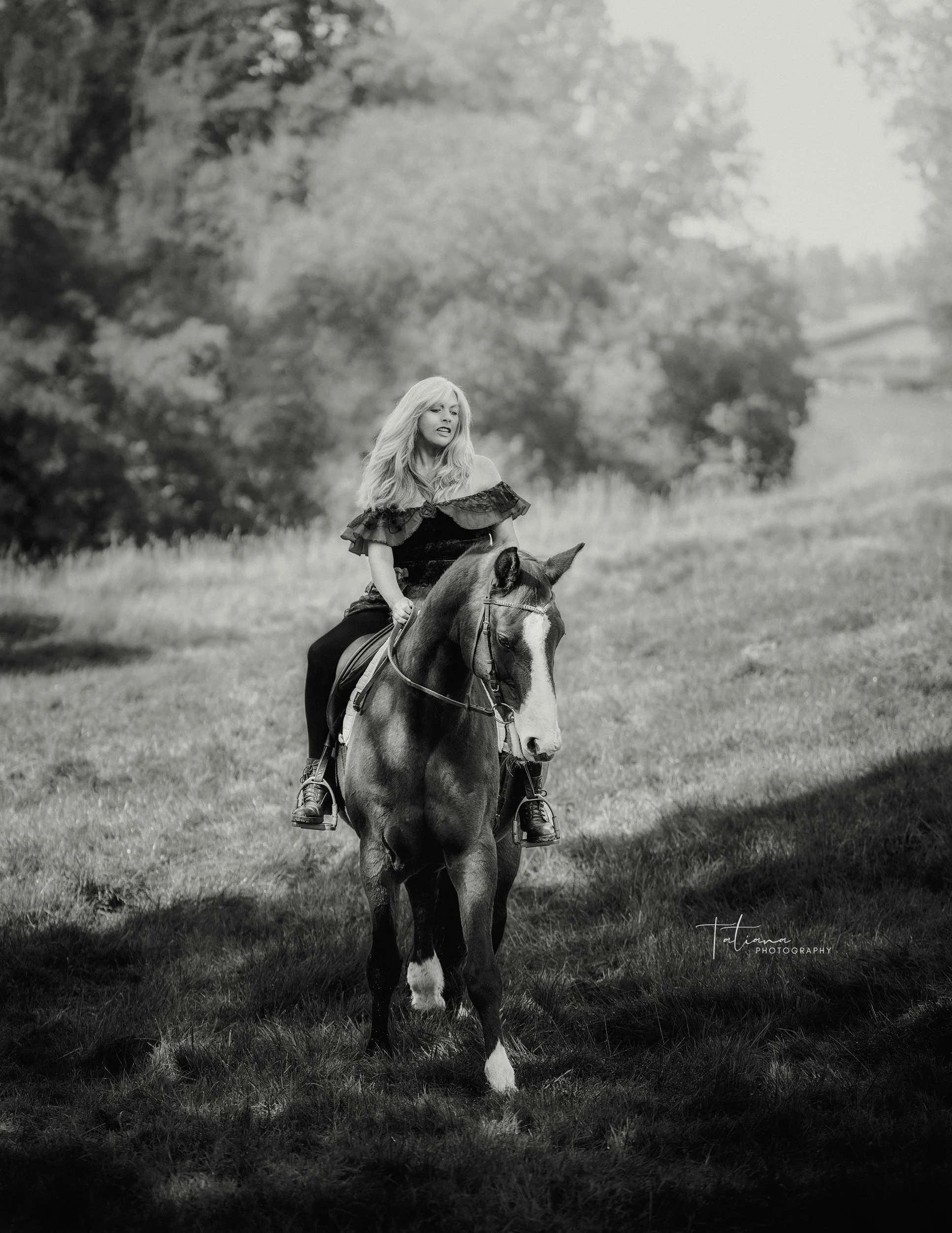 A woman with long blonde hair riding a horse through a grassy field with trees in the background.