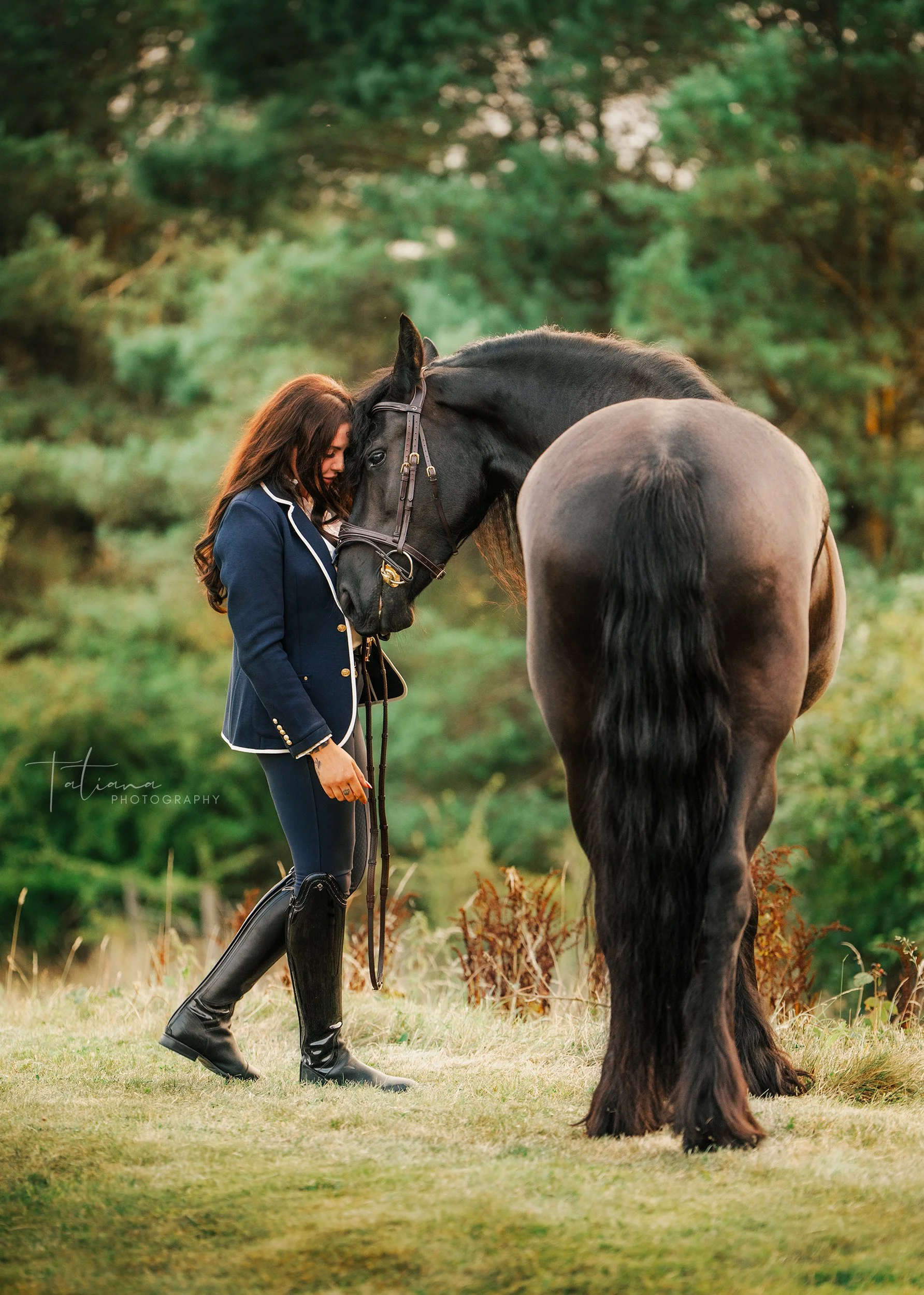 A woman wearing riding attire with tall black riding boots and a navy blazer with white trim stands outdoors, resting her forehead against the forehead of a large black horse. The background shows green trees and grass, indicating a natural setting.