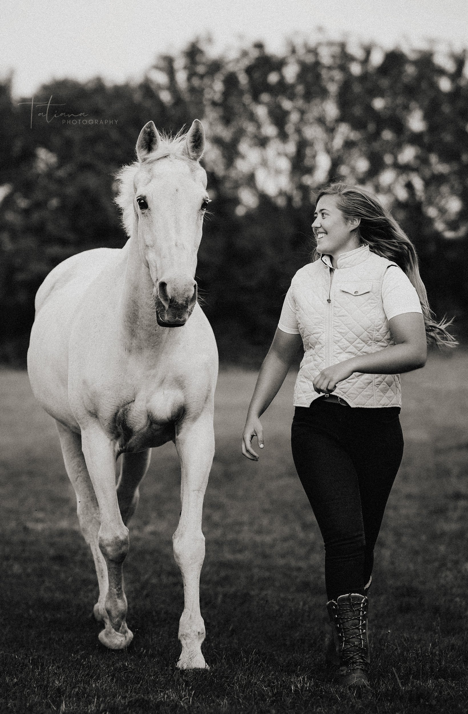 A woman walking with a white horse in an outdoor field, smiling and looking at the horse, black and white photo.