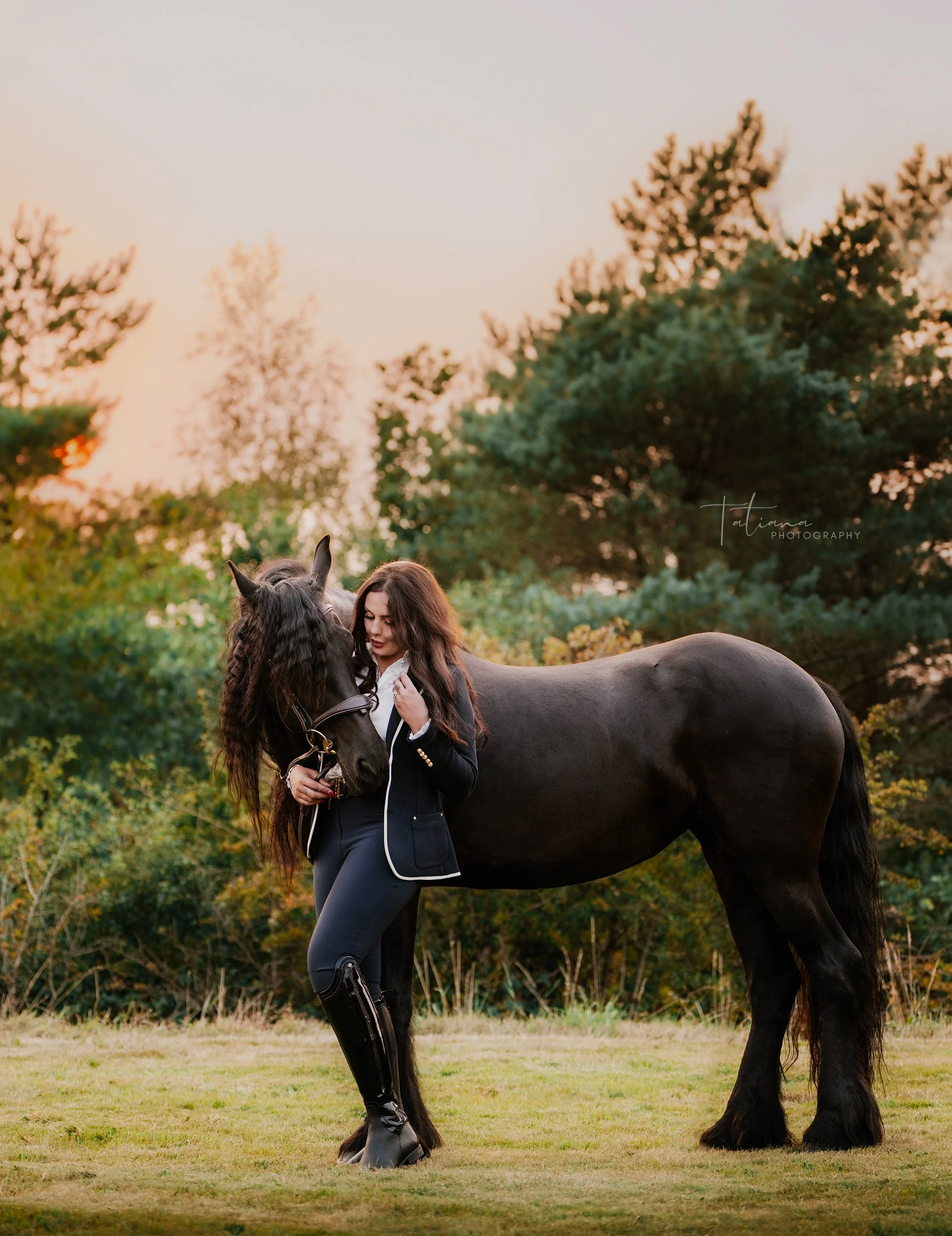 A woman in riding attire embracing a black horse outdoors at sunset, with trees in the background.