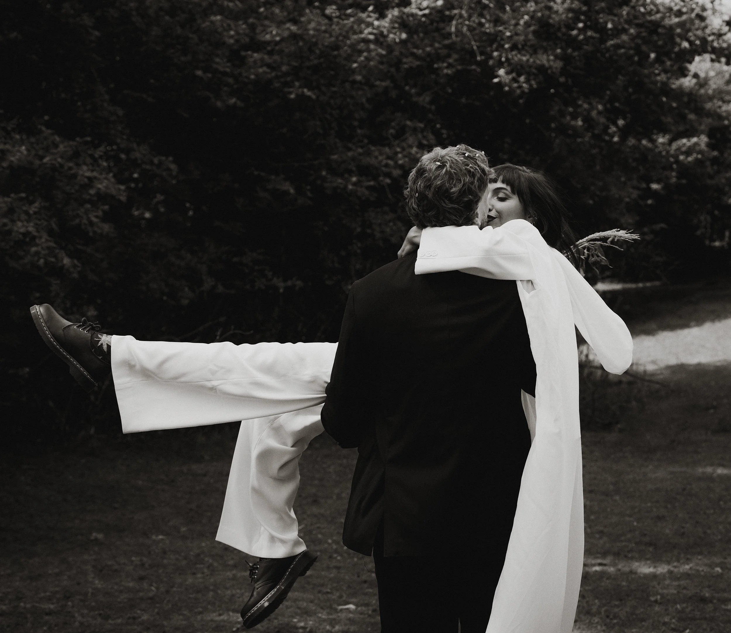 A black and white photo of a couple outdoors, with the man holding the woman in his arms. The woman has her arms around the man's neck, smiling. The woman is wearing a white outfit and the man a dark suit. The background shows trees.