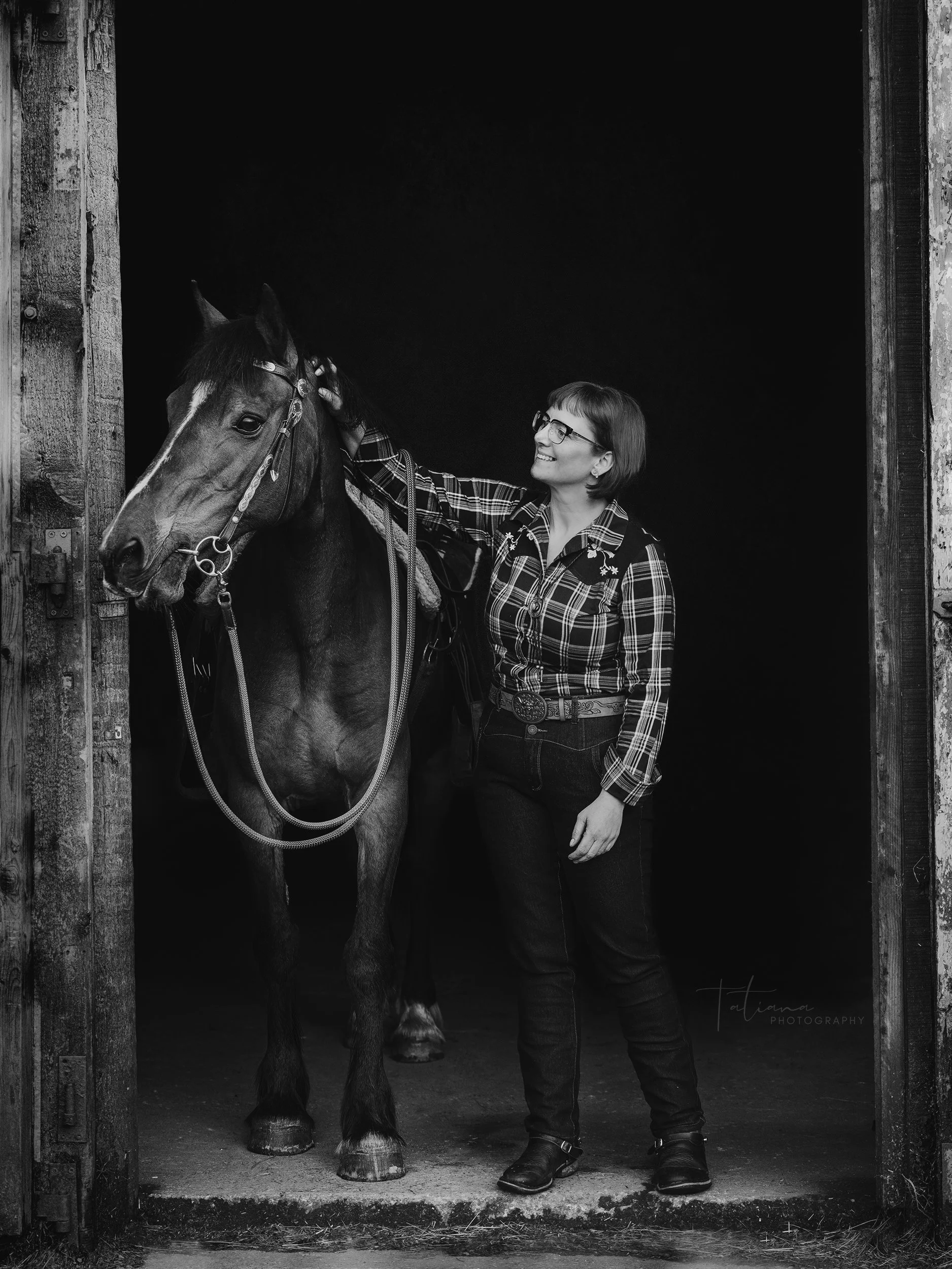 A woman in plaid shirt and jeans standing in a barn doorway, smiling, petting a horse.