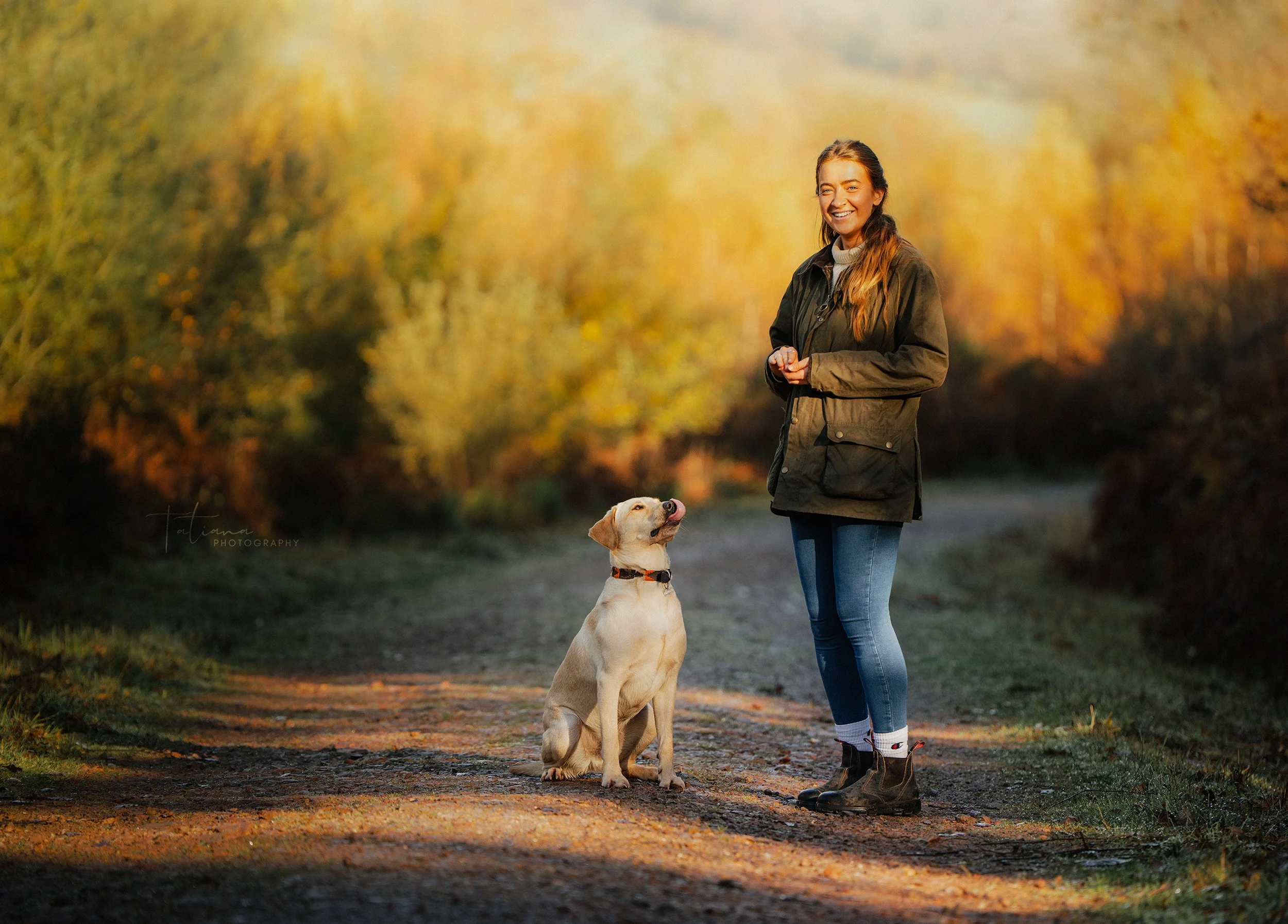 A woman and a Labrador Retriever dog standing on a rural dirt path surrounded by autumn trees with golden leaves. The woman is smiling and wearing a green jacket, jeans, and hiking boots. The dog is sitting and looking up at her, with its tongue slig