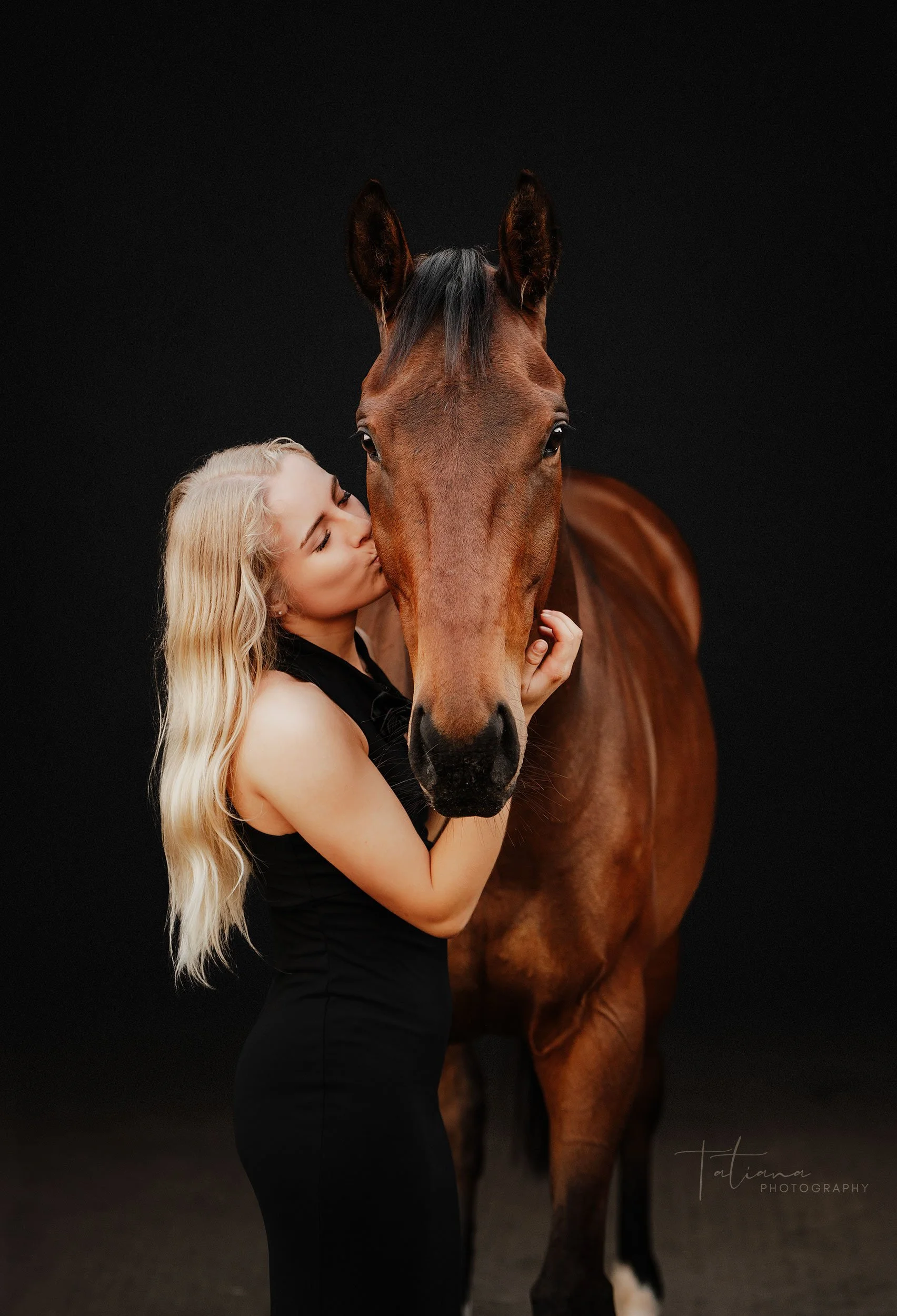 A woman with long blonde hair kissing a brown horse's face while holding its head gently, set against a dark background.